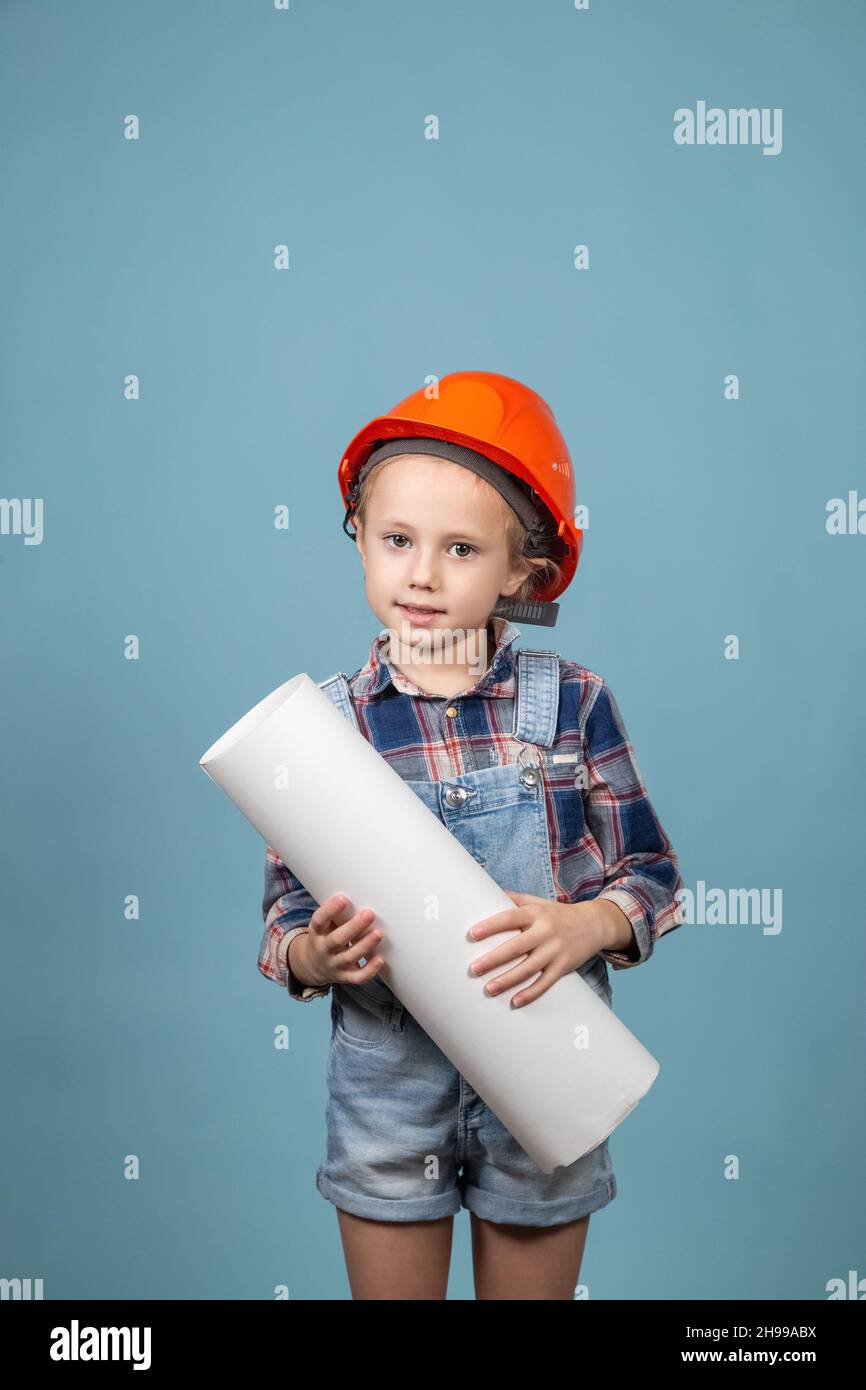 Portrait of cute little caucasian girl builder in hardhats holding construction drawing against blue background. Concept of building a house for a hap Stock Photo