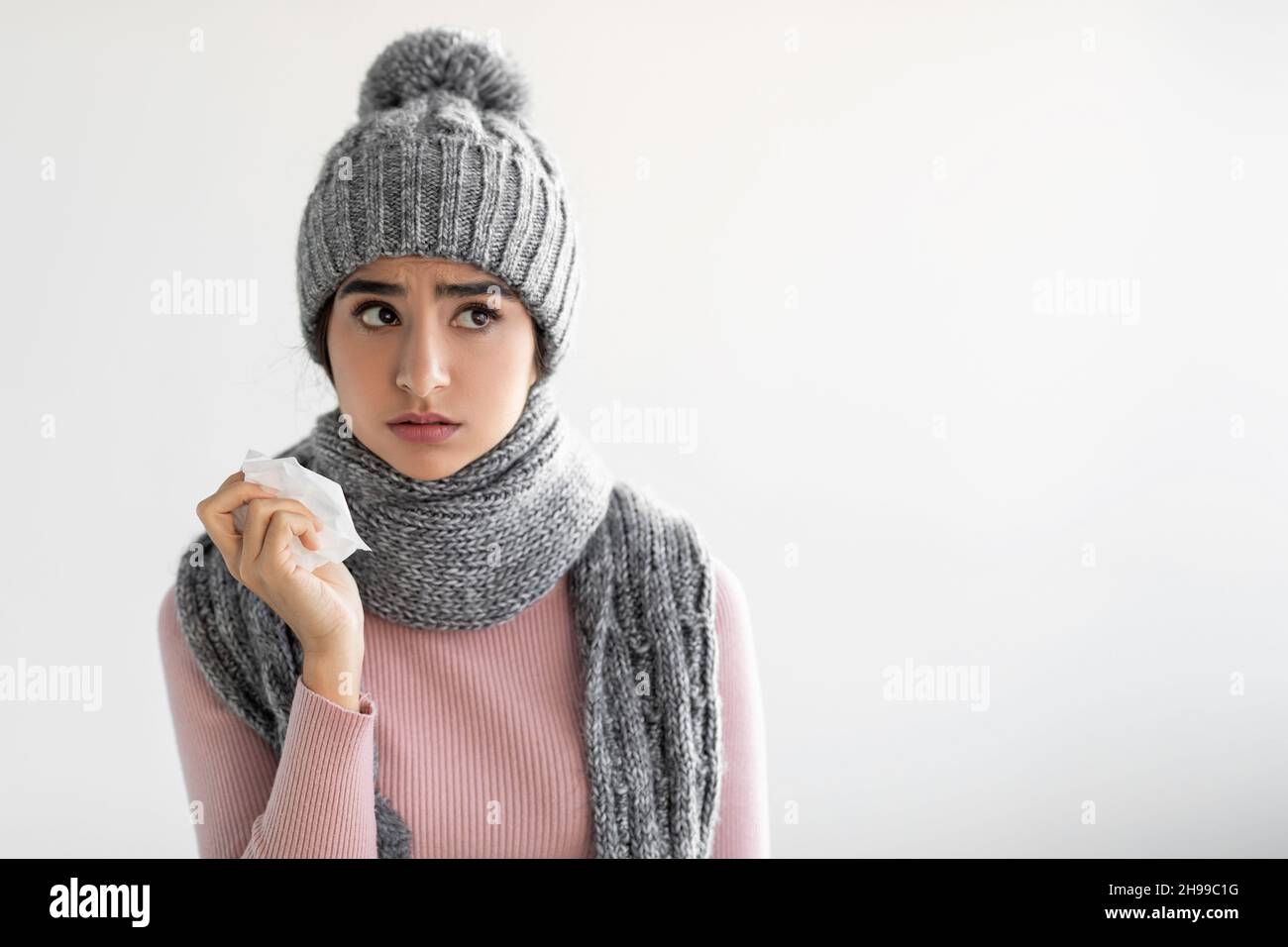 Sad young pretty hindu woman in hat and scarf suffering from flu, holds napkin on gray wall background Stock Photo