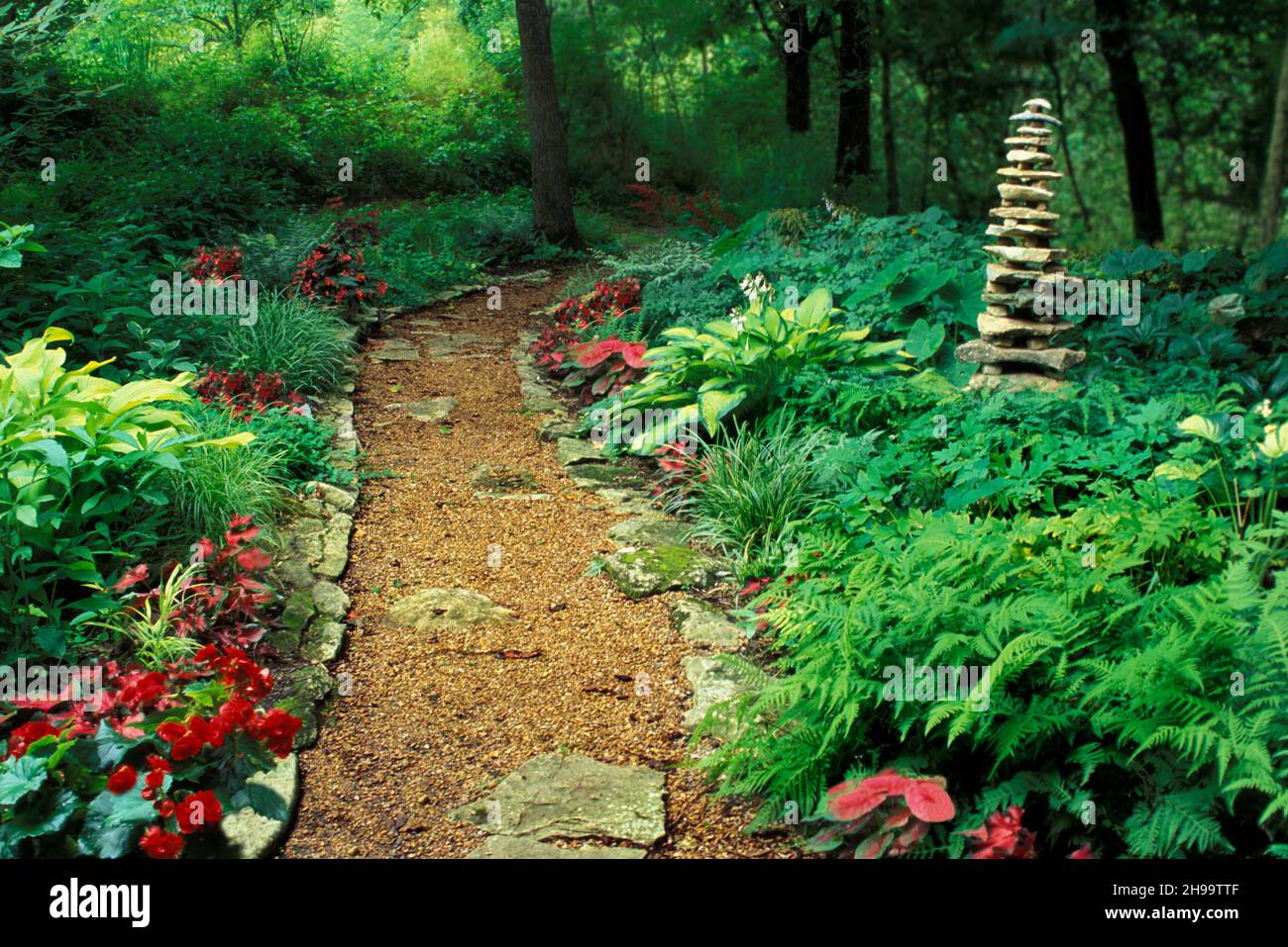 Beautiful hand made rock cairn stands in a lush shade garden with a gravel walkway through ferns and blooming flowers, Missouri, USA Stock Photo
