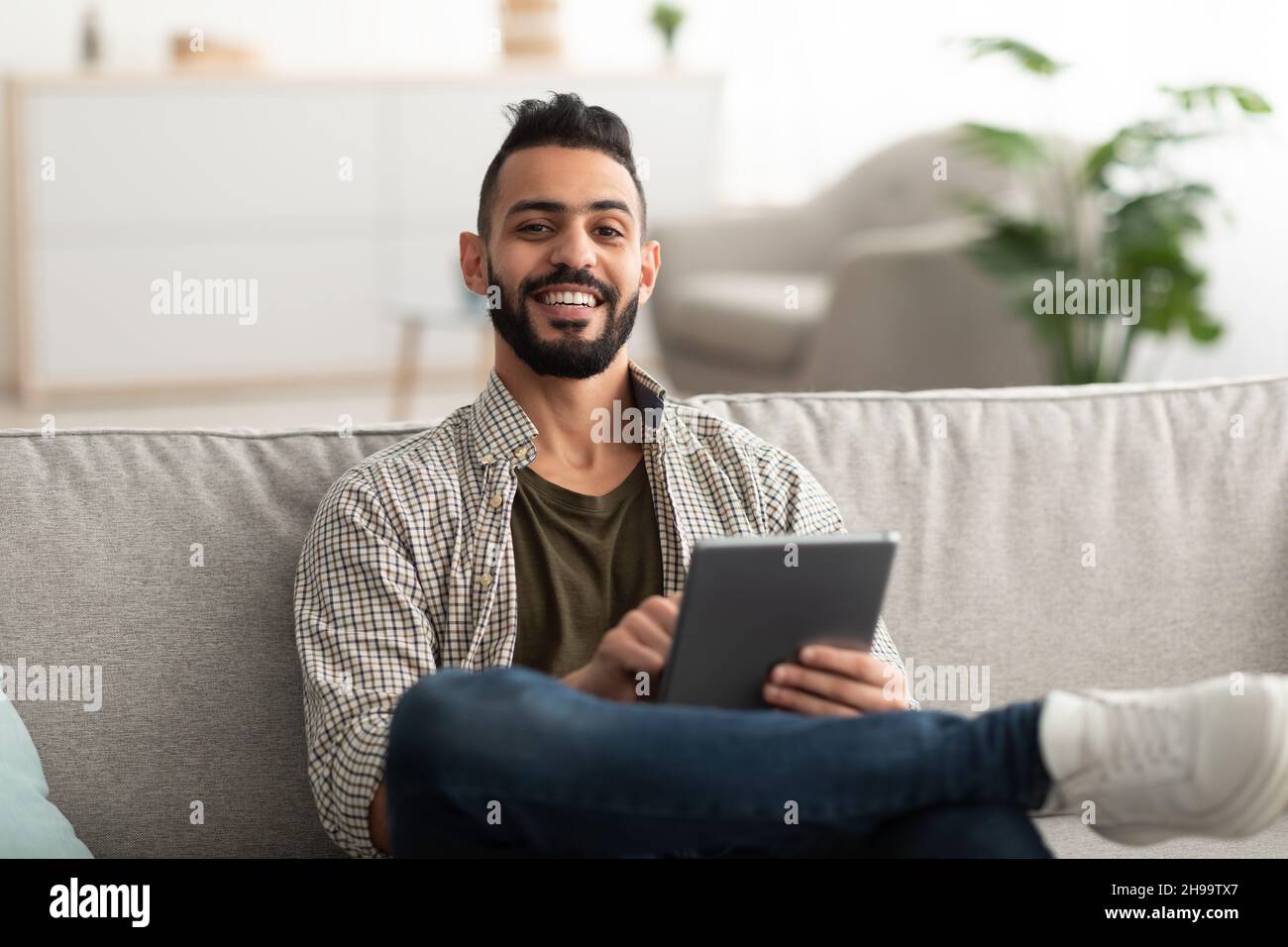 Handsome young Arab man using digital tablet while sitting on couch at home Stock Photo