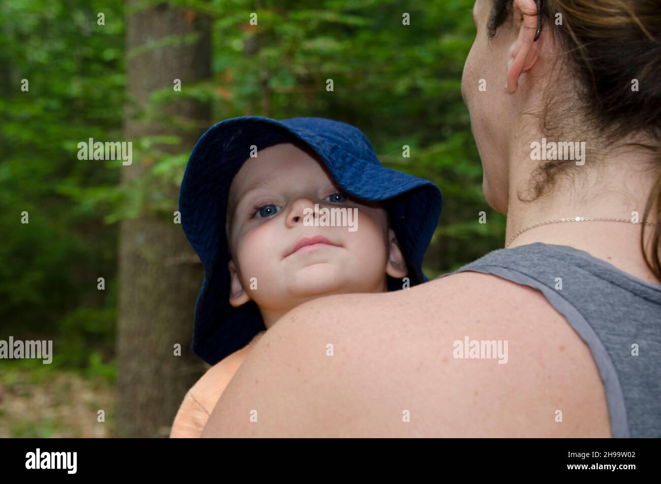 Infant boy with floppy blue cotton hat looking over behind his mother's shoulder in  the garden, Maine, USA Stock Photo