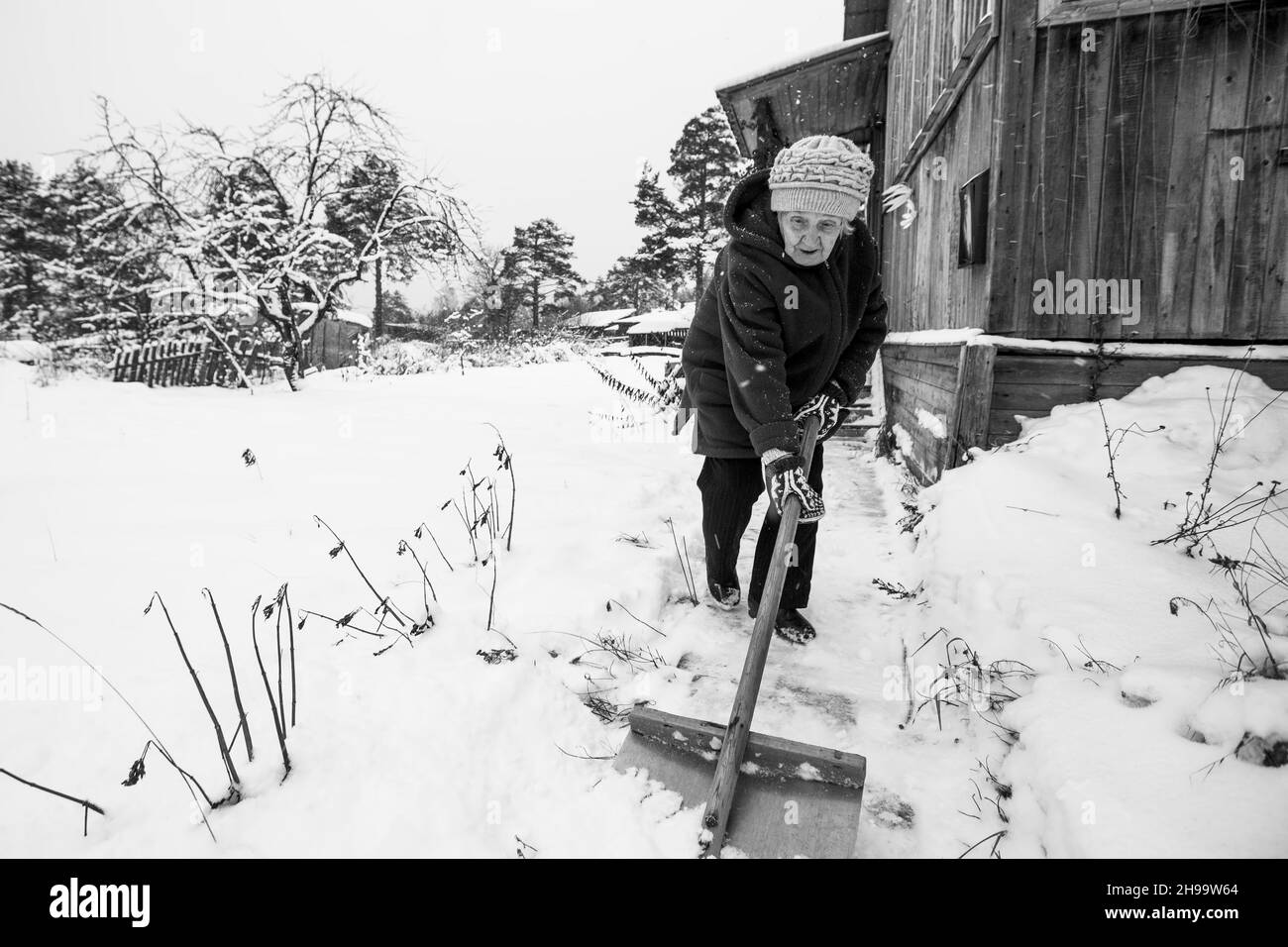 An old woman is shoveling snow outside her rural home. Black and white