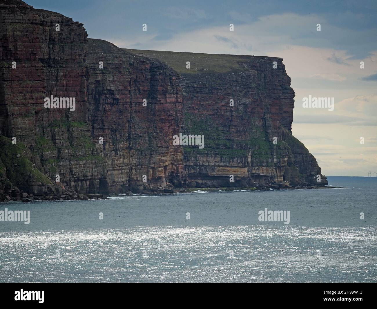 Towering rocky sea-cliffs above Rackwick Bay with silver light on the glittering sea and distant wind turbines - Isle of Hoy, Orkney, Scotland, UK Stock Photo