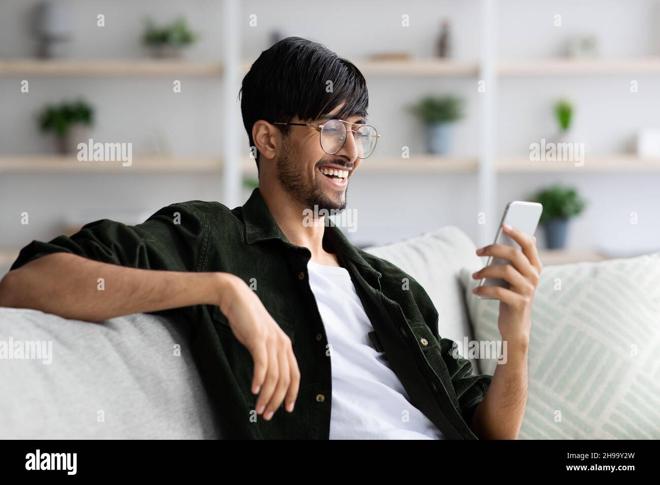Happy indian guy sitting on couch, using smartphone Stock Photo