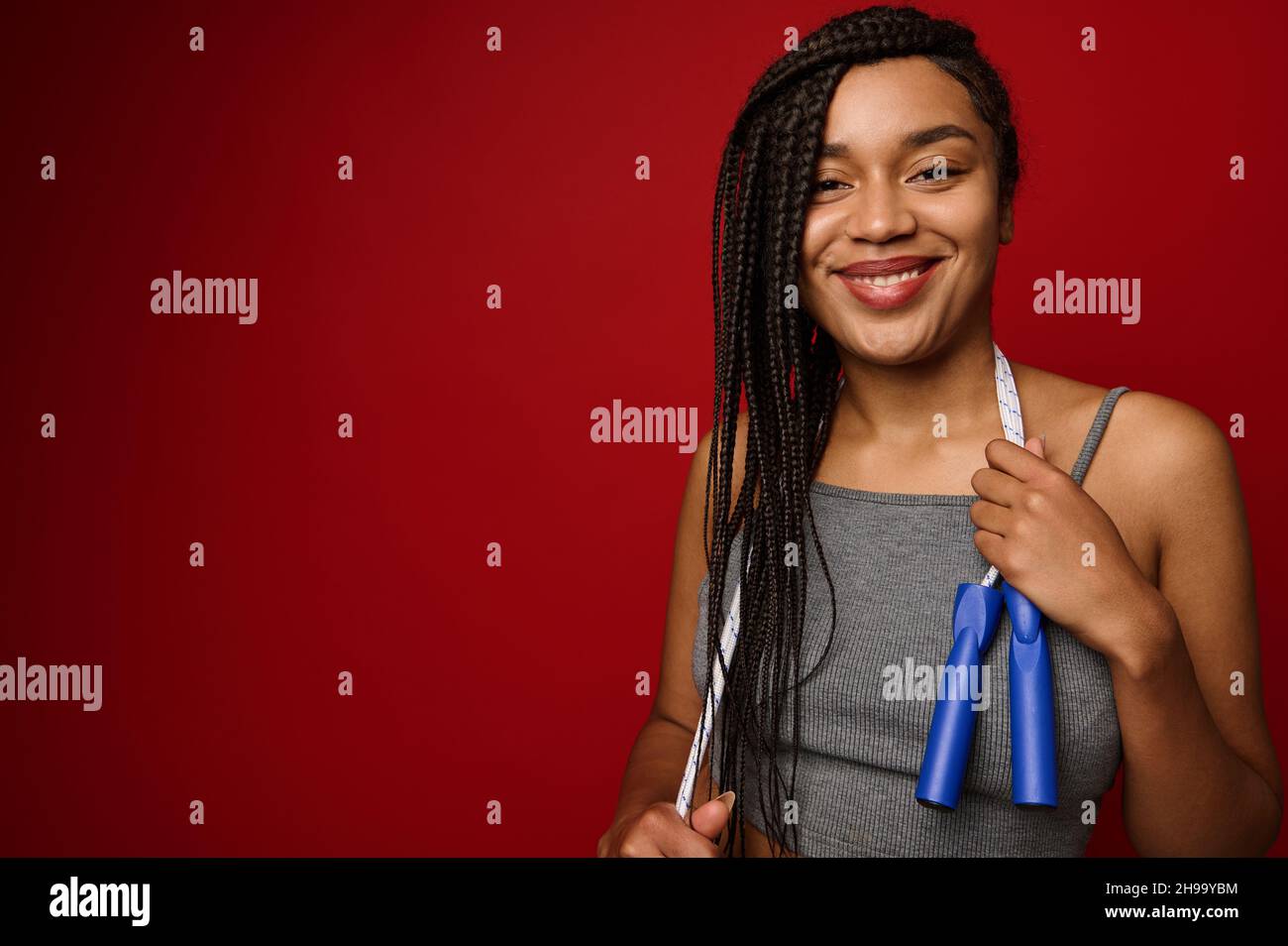 Cheerful African woman with dreadlocks posing against a red background with a skipping rope around her neck, smiling with a toothy smile, looking at t Stock Photo