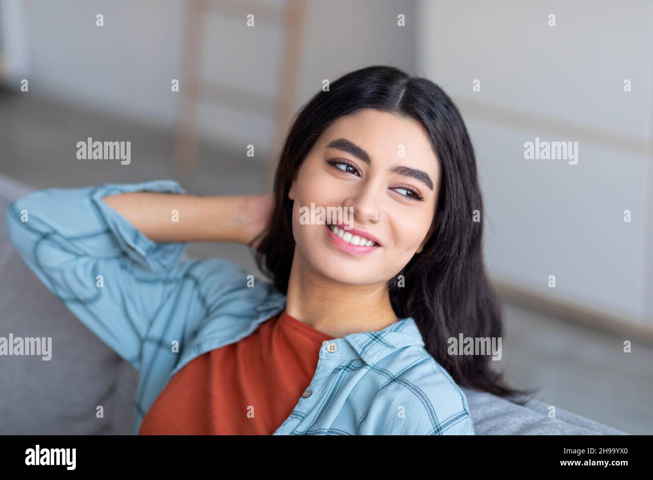 Happy Arab woman leaning back on sofa, resting on comfy couch, holding one hand behind her head, enjoying relaxing day Stock Photo