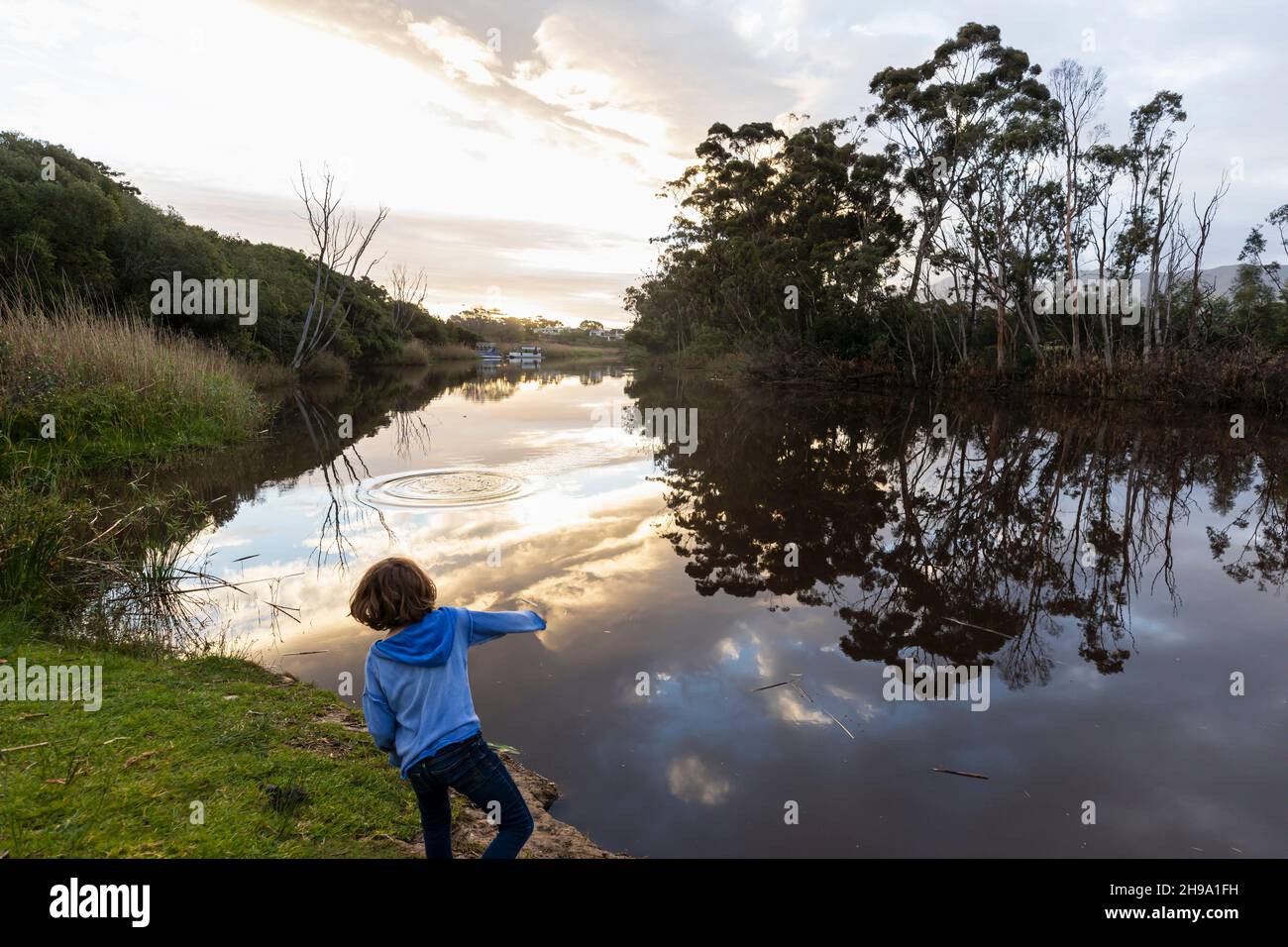 A young boy standing by a river at dusk, sky reflections in the flat calm water Stock Photo