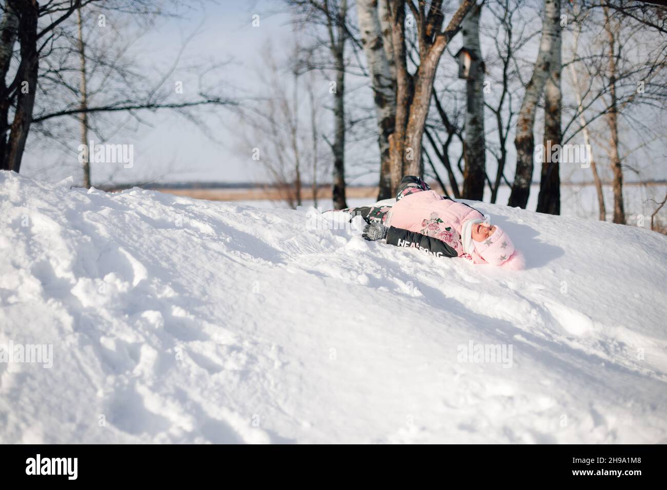 Child rides on snow slide. Girl enjoys winter walk in nature in forest, has fun, plays and lies on snow Stock Photo