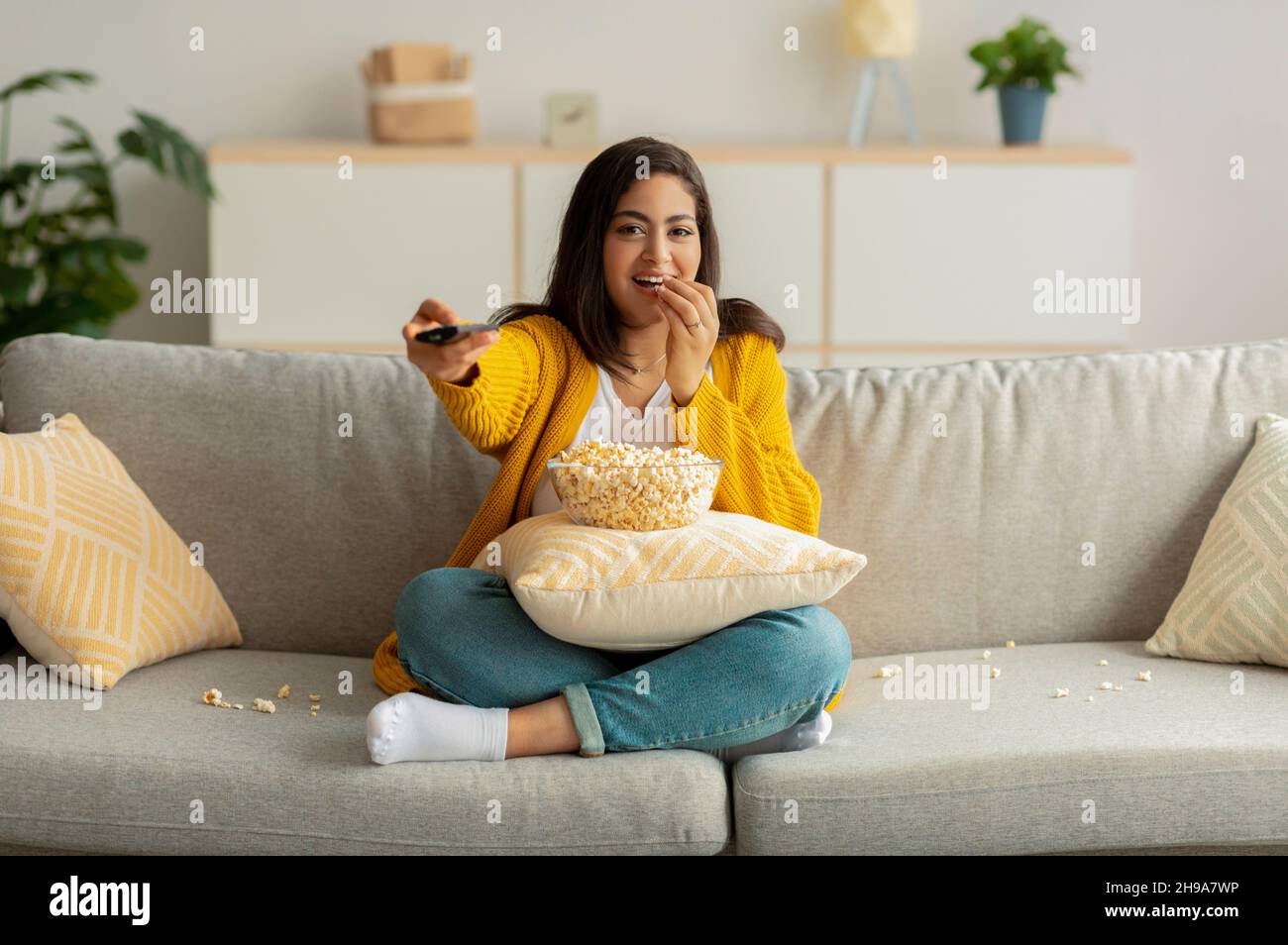 Young arab woman sitting on couch with TV remote, choosing movie to watch and eating popcorn, enjoying free time Stock Photo