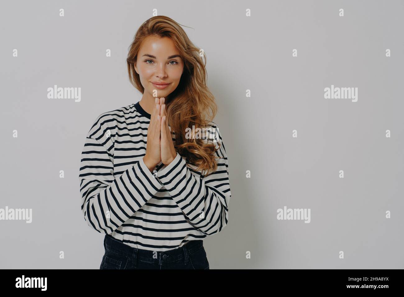 Pleading young blonde woman girl in casual striped shirt holding hands folded in praying gesture Stock Photo