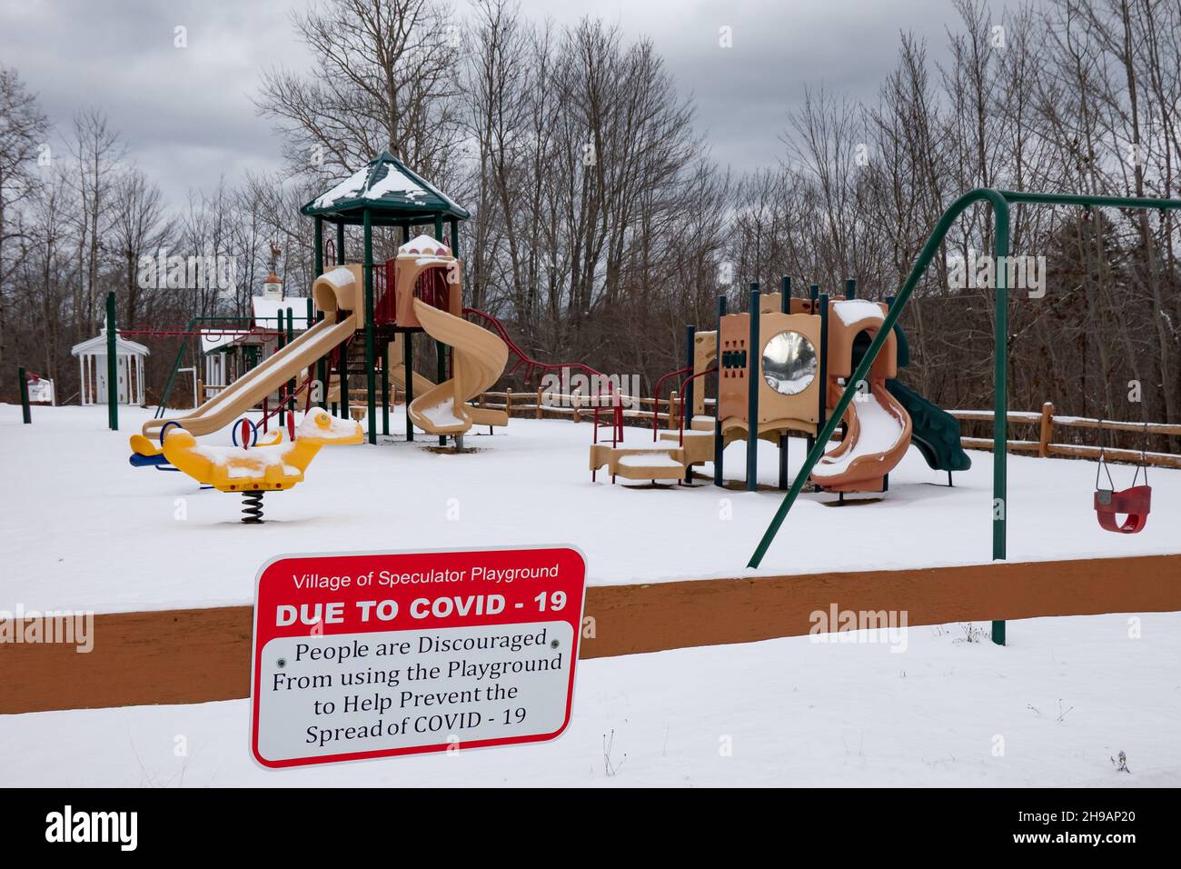 An empty snow covered playground in Speculator, NY USA with a COVID-19 warning sign discouraging use. Stock Photo