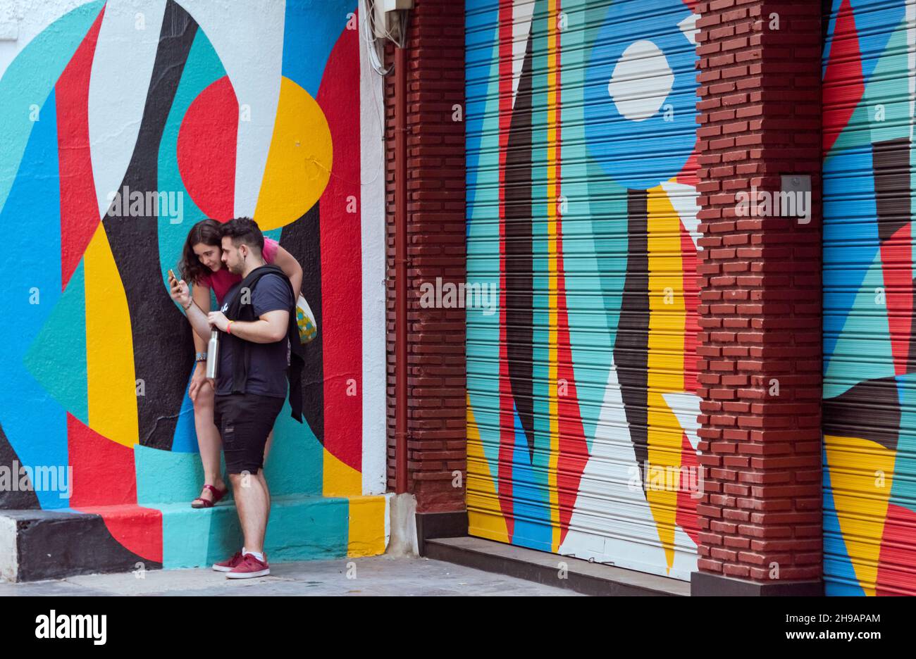 A couple looking at cell phone on the street in front of colorful mural, Valencia, Spain Stock Photo