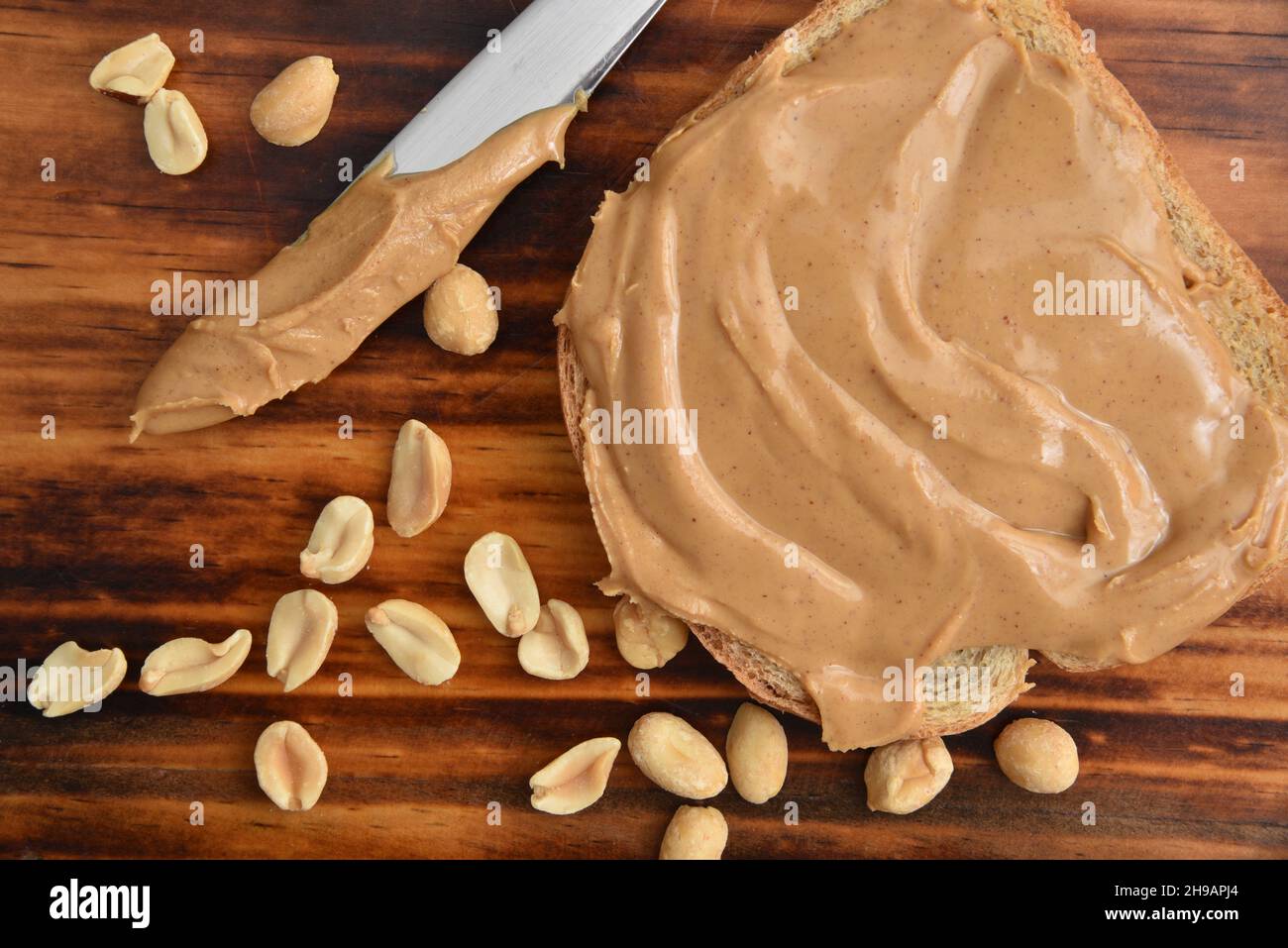 Peanut butter on a slice of bread on a wooden table with roasted salted peanuts overhead view Stock Photo