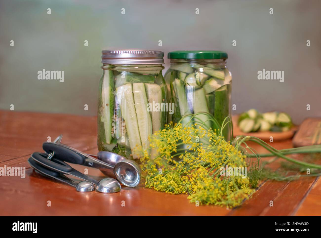 Jars of homemade pickles sit with fresh dill, measuring spoons, sliced