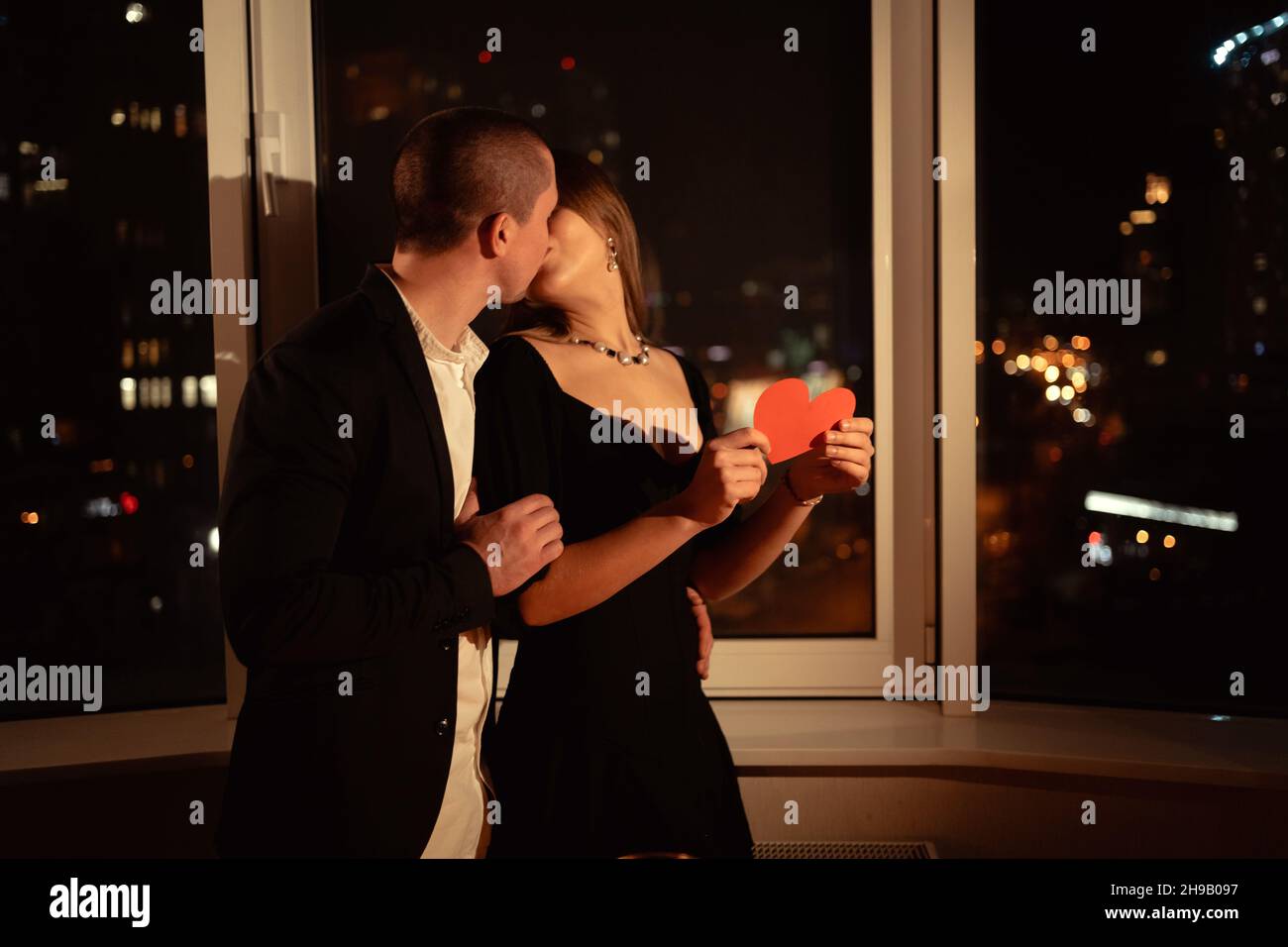 married couple kissing passionately while standing by the window in the evening, celebrating valentine's day, a man kissing a woman on a date Stock Photo
