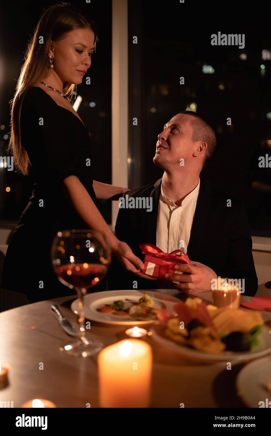 Couple in love on a date valentines day, woman giving a man a gift in a box, romantic date for an anniversary in a restaurant, night candles and wine Stock Photo