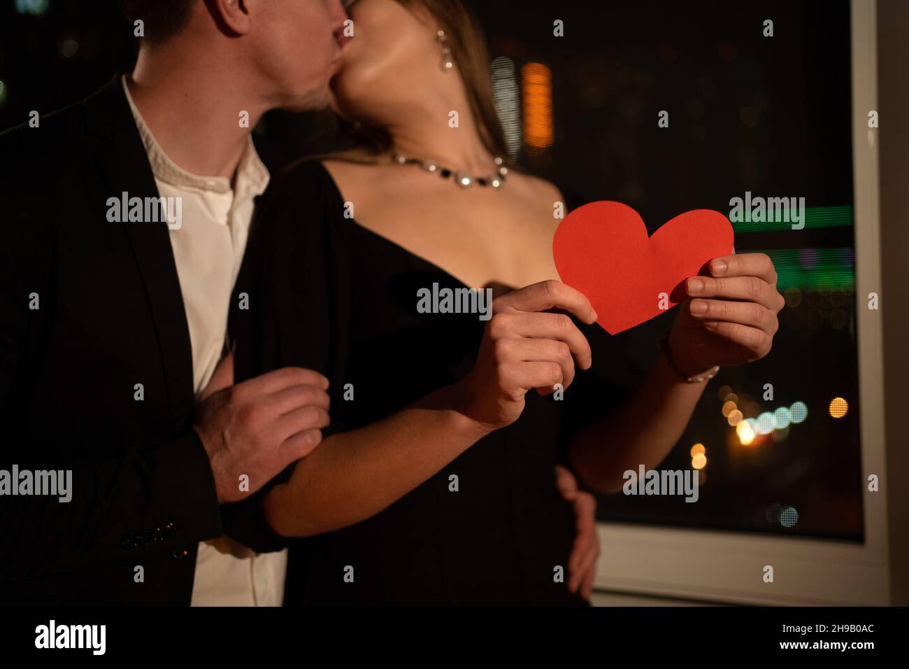 valentine's day, passionate kiss in the evening by the window, night date in the apartment, man kissing a woman, red heart in female hands, close-up Stock Photo