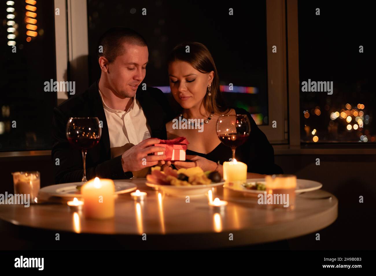 Valentine's day date by candlelight evening, romantic dinner for two, man giving girl an anniversary gift at the table, man and woman relationship Stock Photo