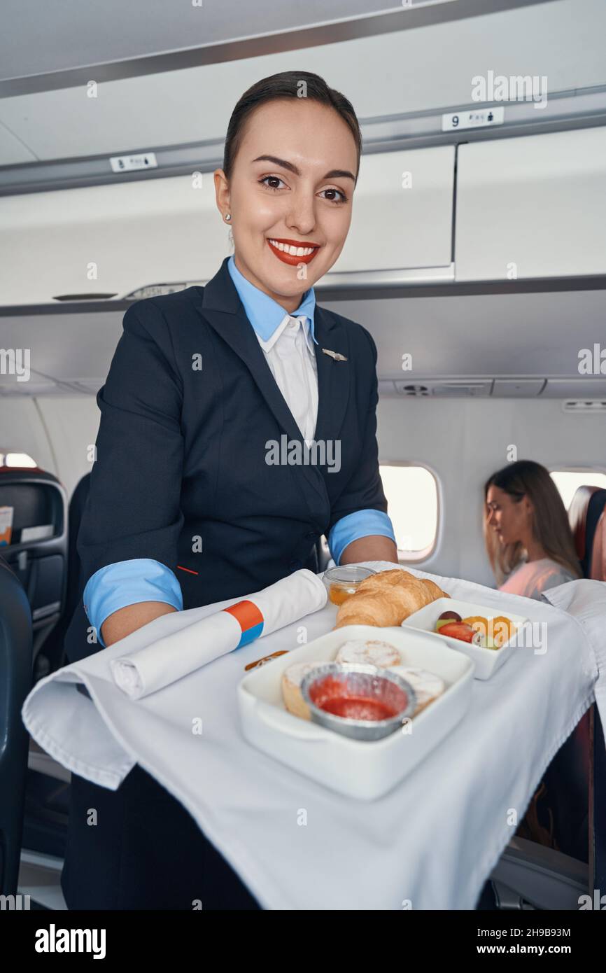 Joyful stewardess rolling airline meal trolley through aisle Stock