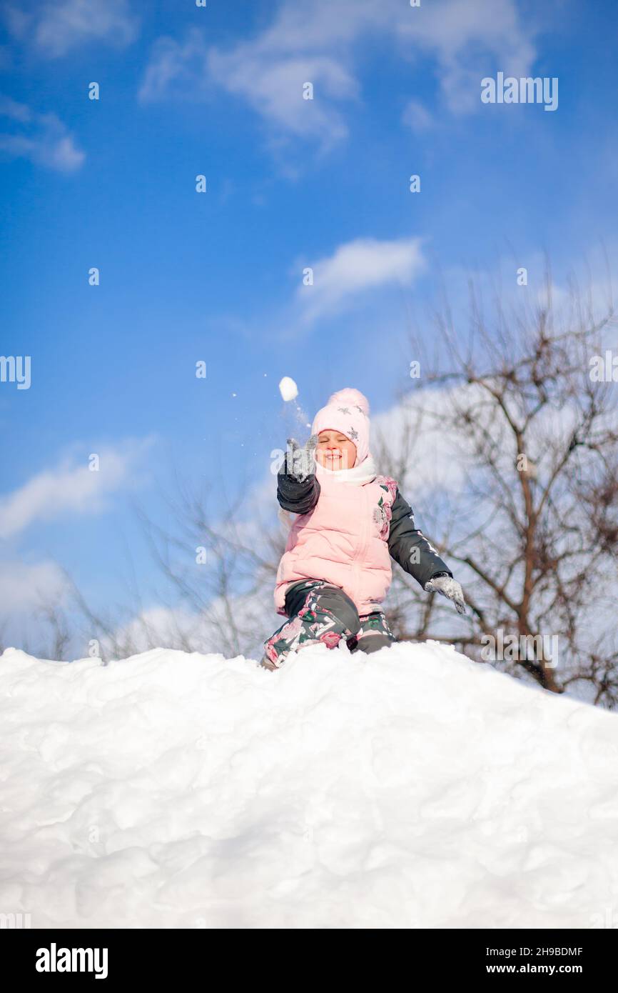 Little girl is playing snowballs. Child has fun on sunny winter day, plays and rides snow slide without sled and jumps into snowdrifts, winter Stock Photo