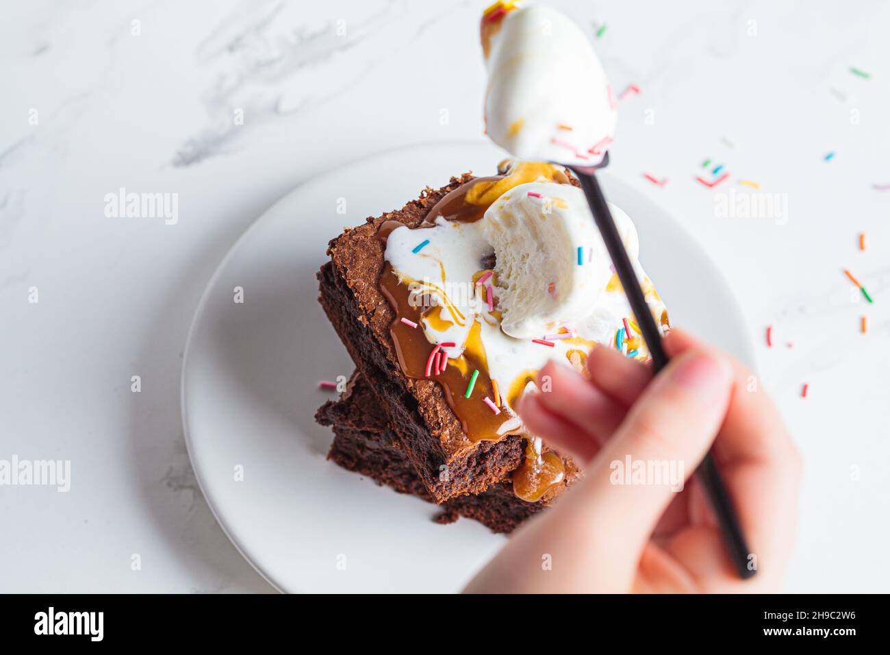 Children's hand eating brownie with a scoop of ice cream, caramel and sprinkles, white background. Stock Photo