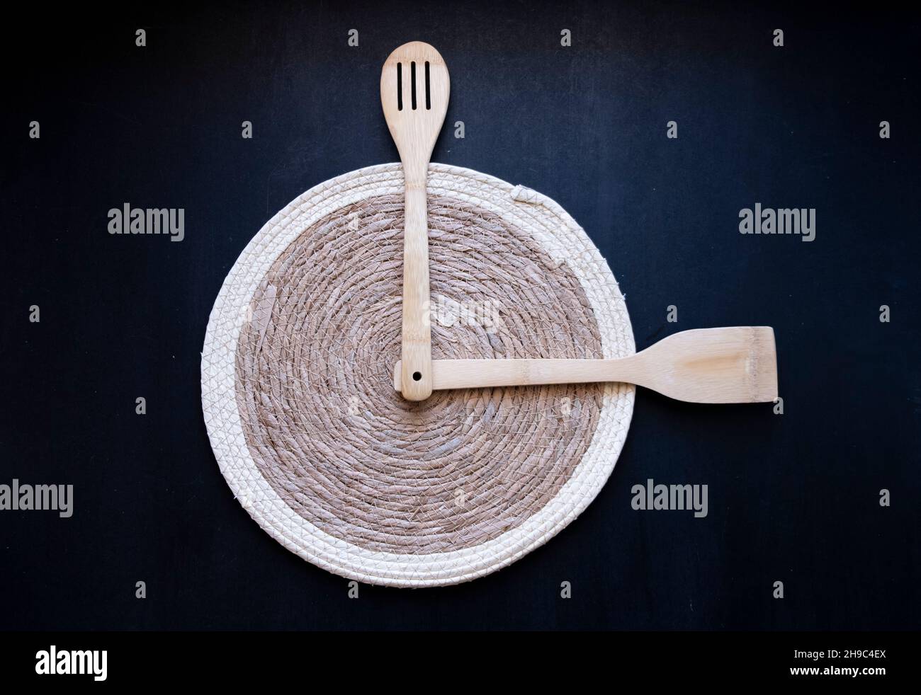 wooden spoon and spatula clock idea on a bamboo serving with, dinner, lunch or breakfast  time, Straw plaid placemat on isolated black background Stock Photo