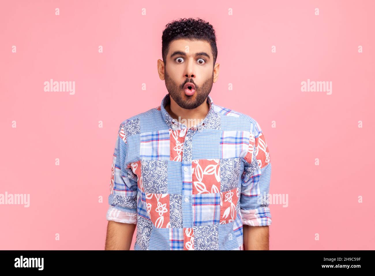 Portrait of excited funny man in blue casual style shirt with pout lips and big amazed eyes, looking surprised and silly at camera, wondered expression. Indoor studio shot isolated on pink background. Stock Photo