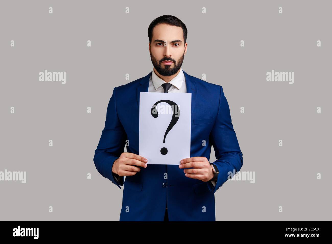 Portrait of serious bearded businessman looking at camera, holding paper with question mark, thinks about tasks, wearing official style suit. Indoor studio shot isolated on gray background. Stock Photo