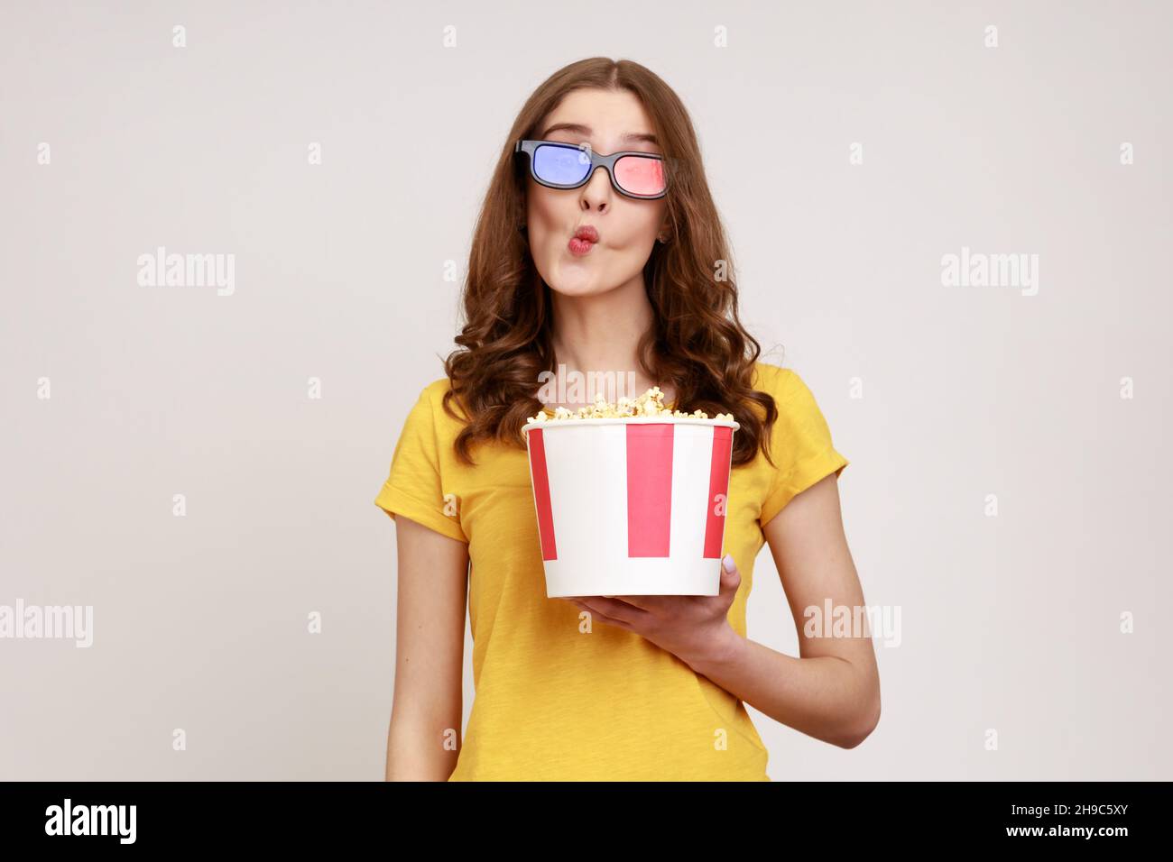 Young childish woman in 3d glasses and yellow casual t-shirt, watching movie film, holding bucket of popcorn, making fish face grimace with pout lips. Indoor studio shot isolated on gray background. Stock Photo
