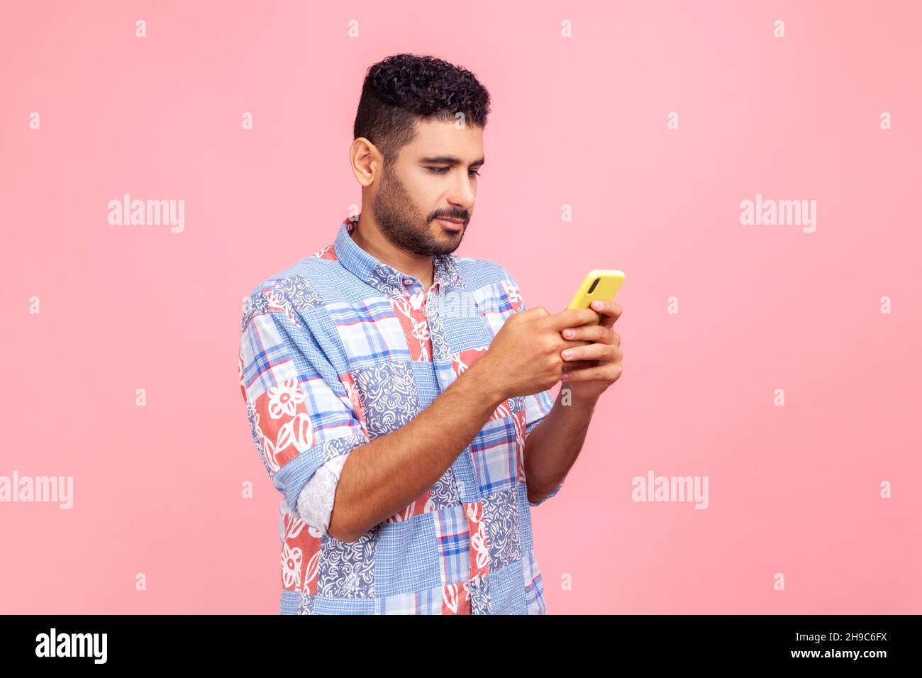 Side view of serious concentrated brunette man with beard holding cell phone in hands, chatting or checking e-mail, gadget addiction. Indoor studio shot isolated on pink background. Stock Photo