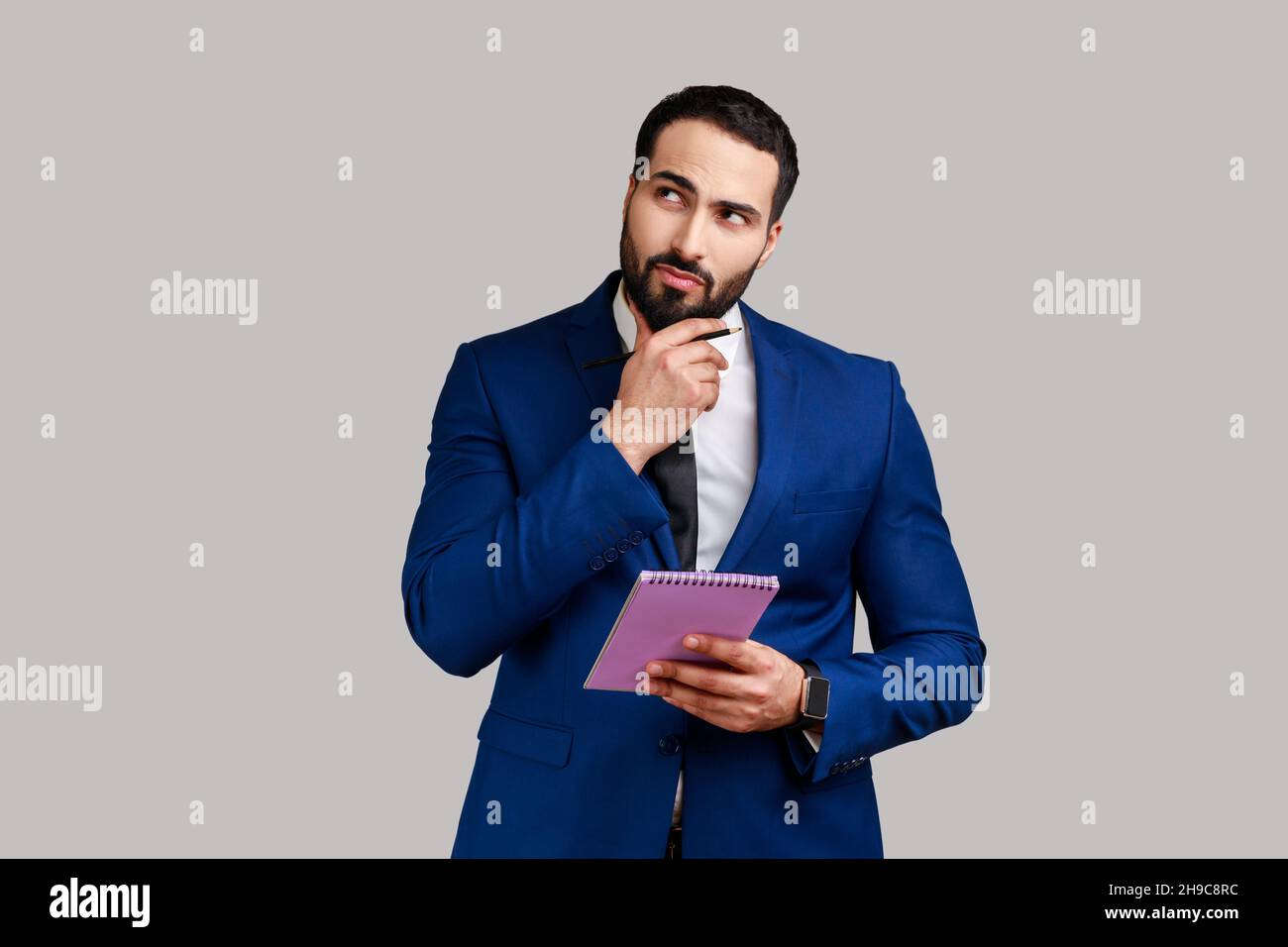 Young adult businessman standing with thoughtful expression, holding paper notebook, pondering business idea, future plans, wearing official style suit. Indoor studio shot isolated on gray background. Stock Photo