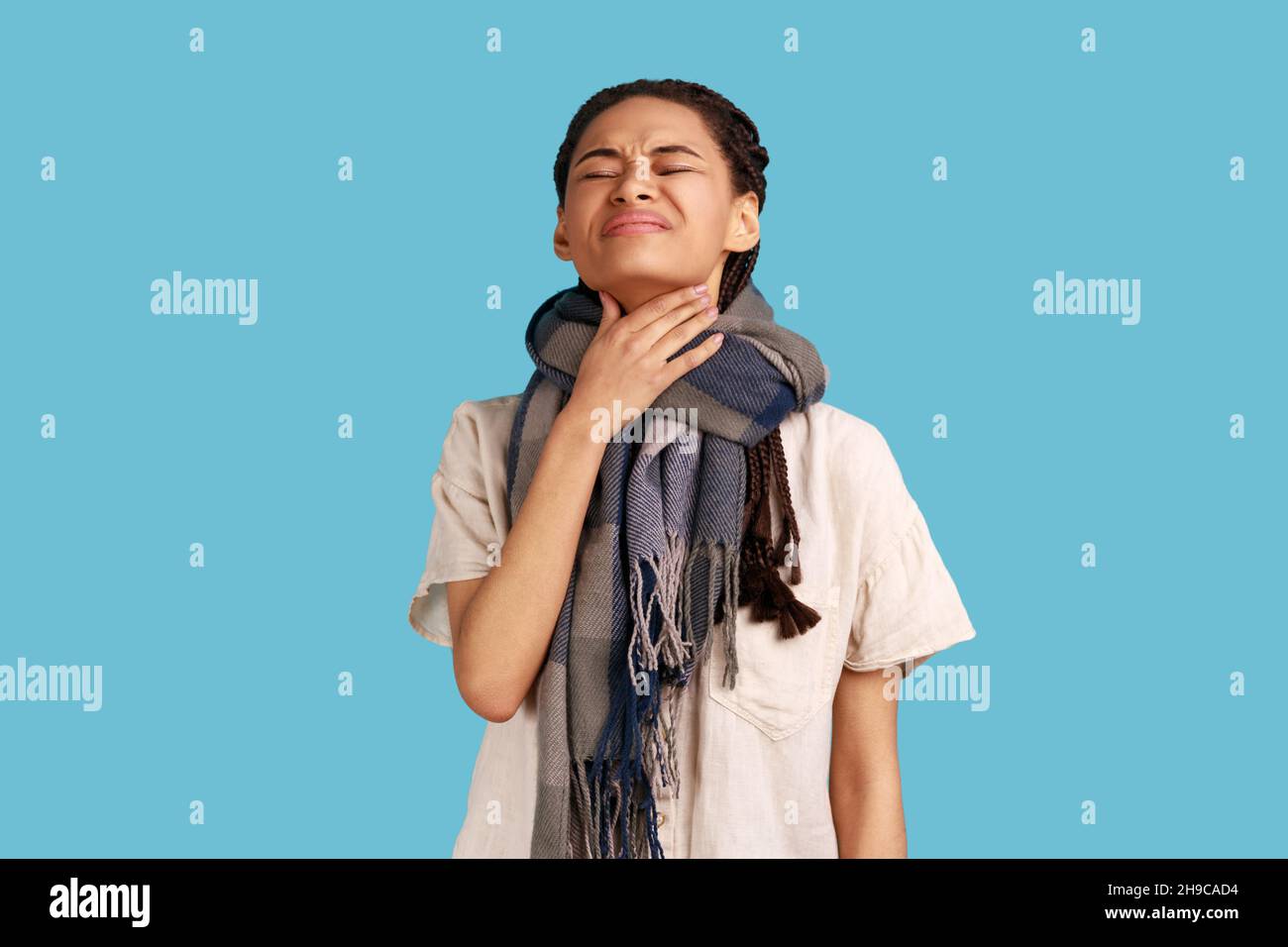 Unhappy sick woman wrapped in scarf, touches neck, suffers from sore throat, cant breath well, chokes and smirks face, feels discomfort and suffocation. Indoor studio shot isolated on blue background. Stock Photo
