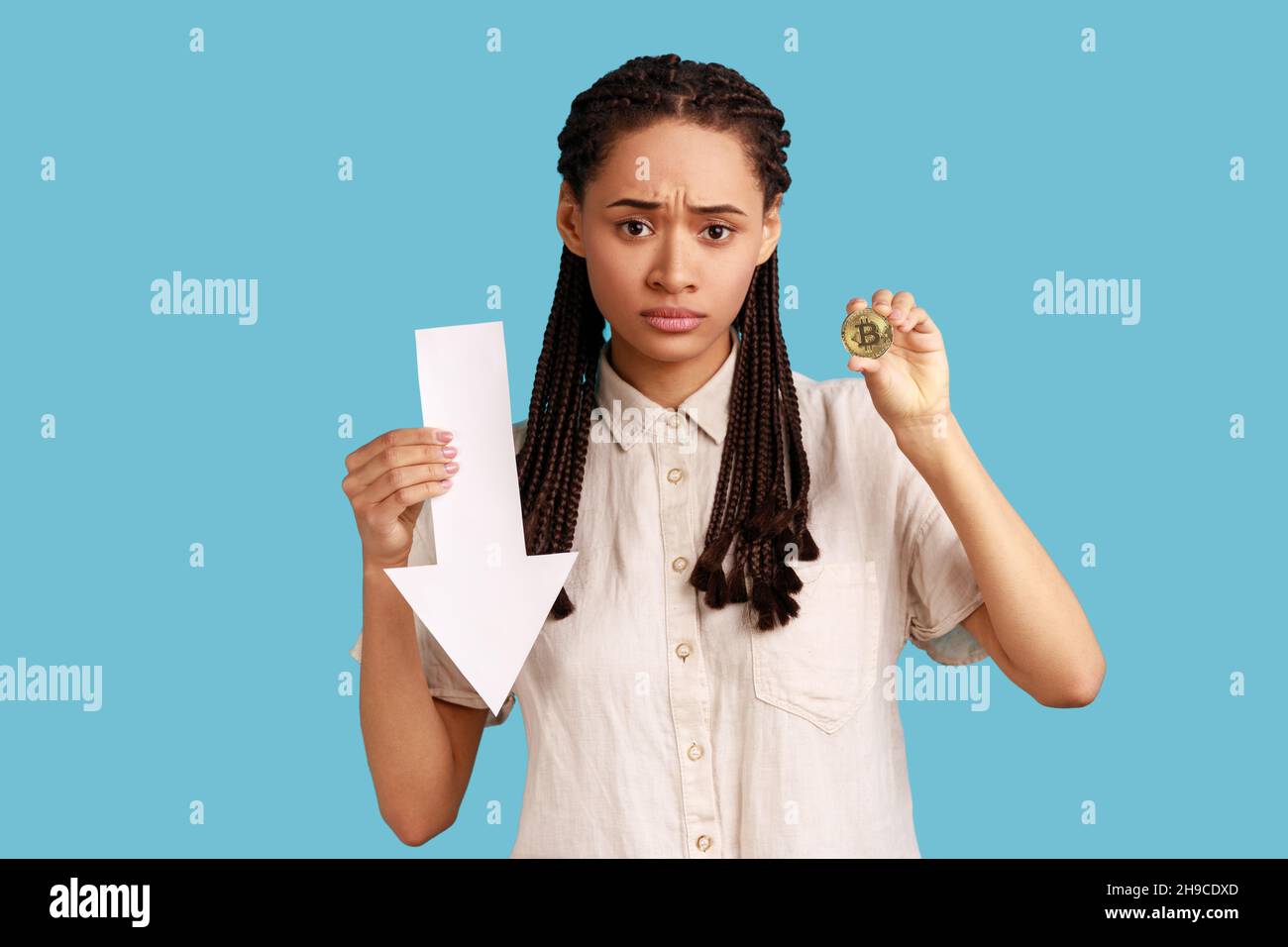 Portrait of dissatisfied woman with dreadlocks showing gold bitcoin and arrow pointing down, downgrade of electronic currency, wearing white shirt. Indoor studio shot isolated on blue background. Stock Photo
