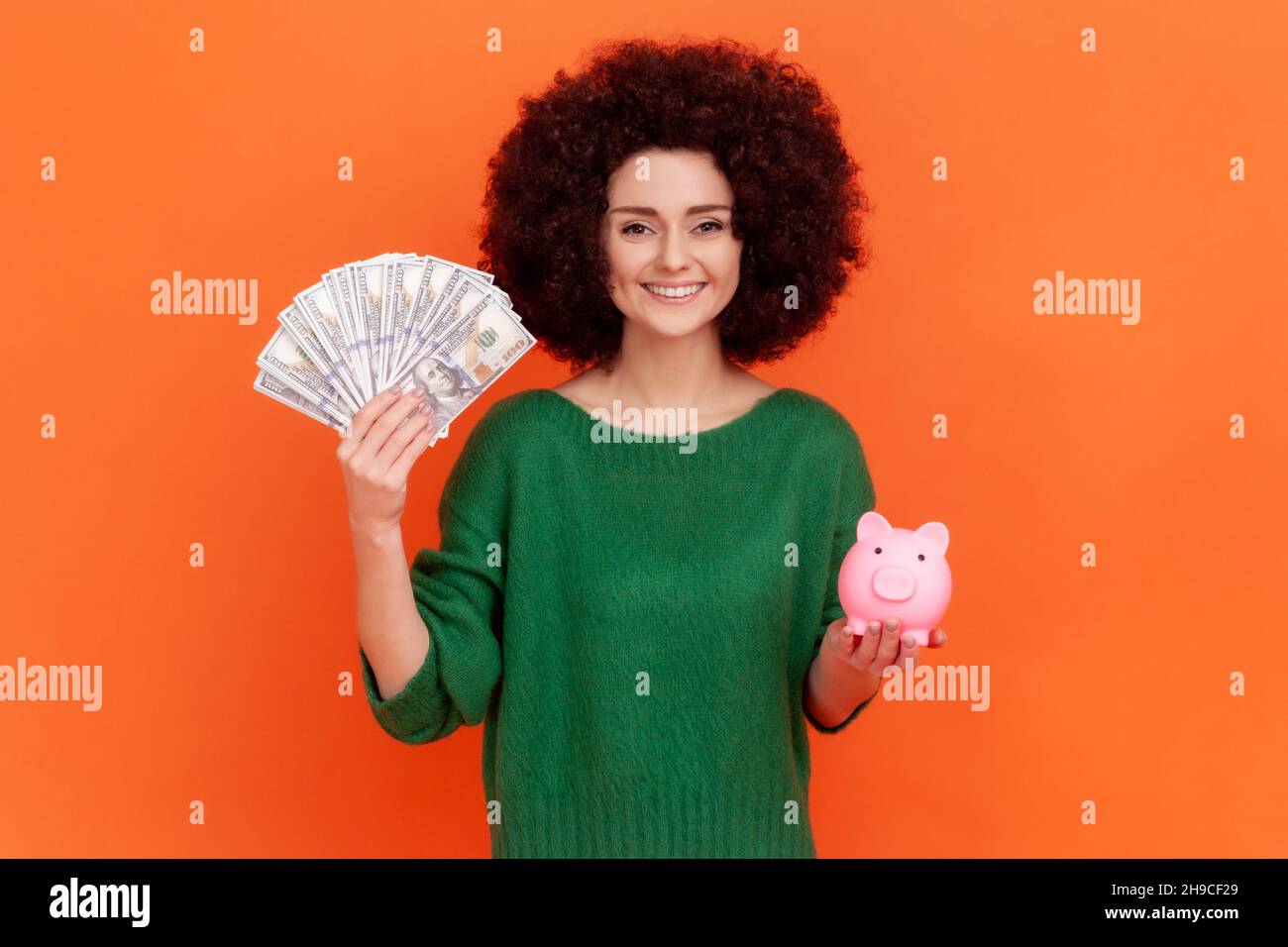Smiling affirmative woman with Afro hairstyle wearing green casual style sweater holding big sum of money and piggy bank, profitable investment. Indoor studio shot isolated on orange background. Stock Photo
