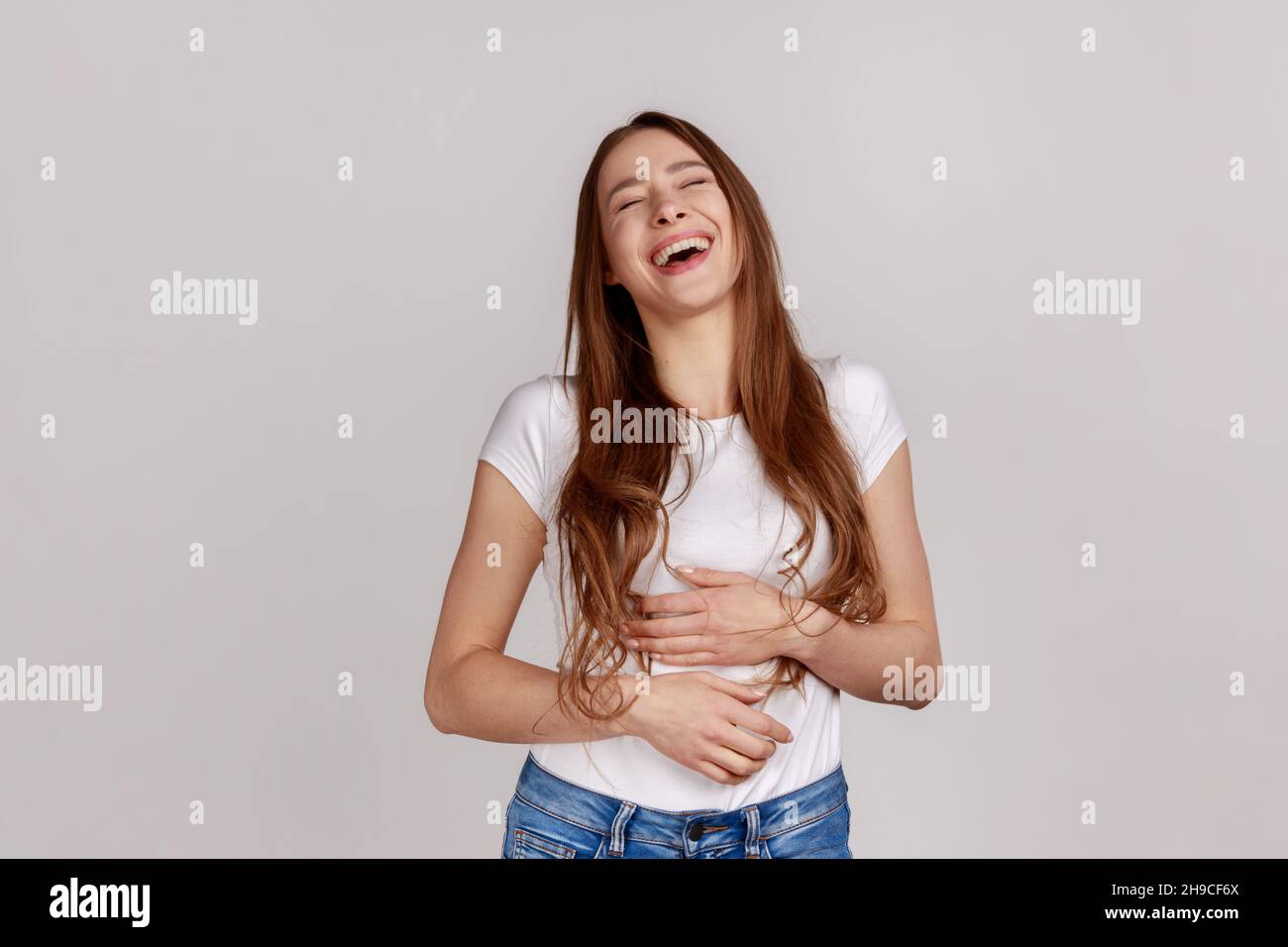 Extremely happy woman holding her stomach and laughing out loud, chuckling giggling at amusing anecdote, sincere emotion, wearing white T-shirt. Indoor studio shot isolated on gray background. Stock Photo