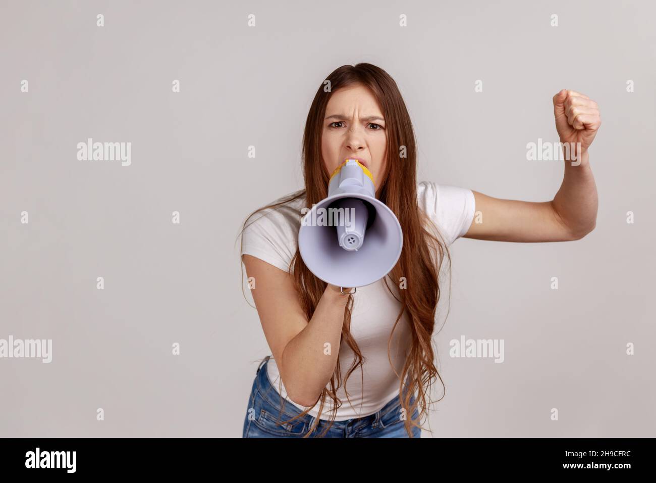 Portrait of woman holding megaphone near mouth, loudly speaking, screaming, making announcement with raised arm, protesting, wearing white T-shirt. Indoor studio shot isolated on gray background. Stock Photo