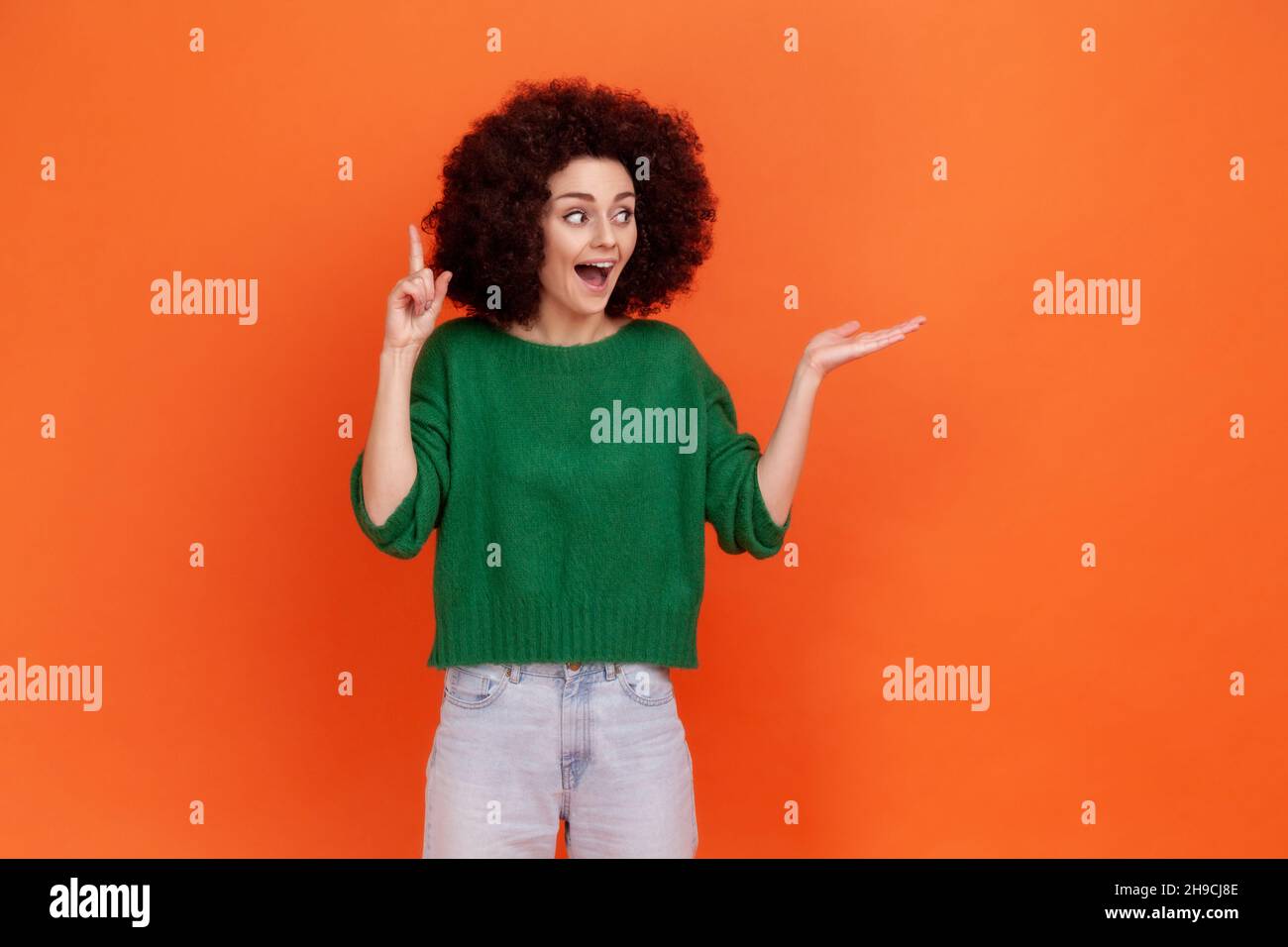 Positive woman with Afro hairstyle wearing green casual style sweater pointing finger up, has idea, presenting area for advertisement. Indoor studio shot isolated on orange background. Stock Photo