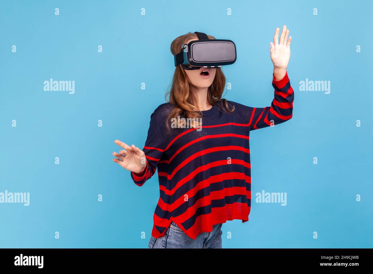 Woman wearing striped casual style sweater and vr headset, playing virtual reality game with shocked face, surprised by innovative technology. Indoor studio shot isolated on blue background. Stock Photo