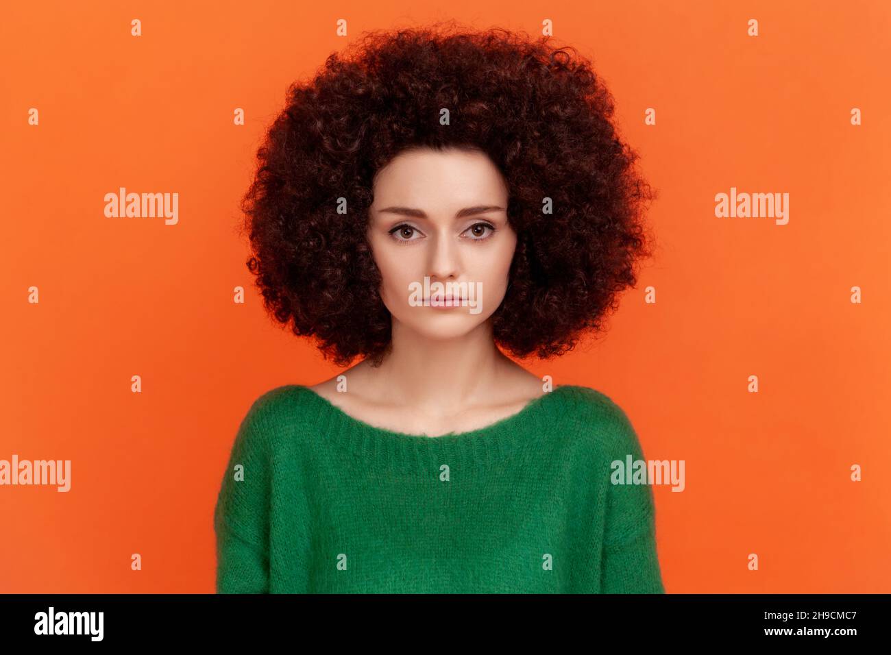 Portrait of serious strict woman with Afro hairstyle wearing green casual style sweater looking at camera with assertive facial expression. Indoor studio shot isolated on orange background. Stock Photo