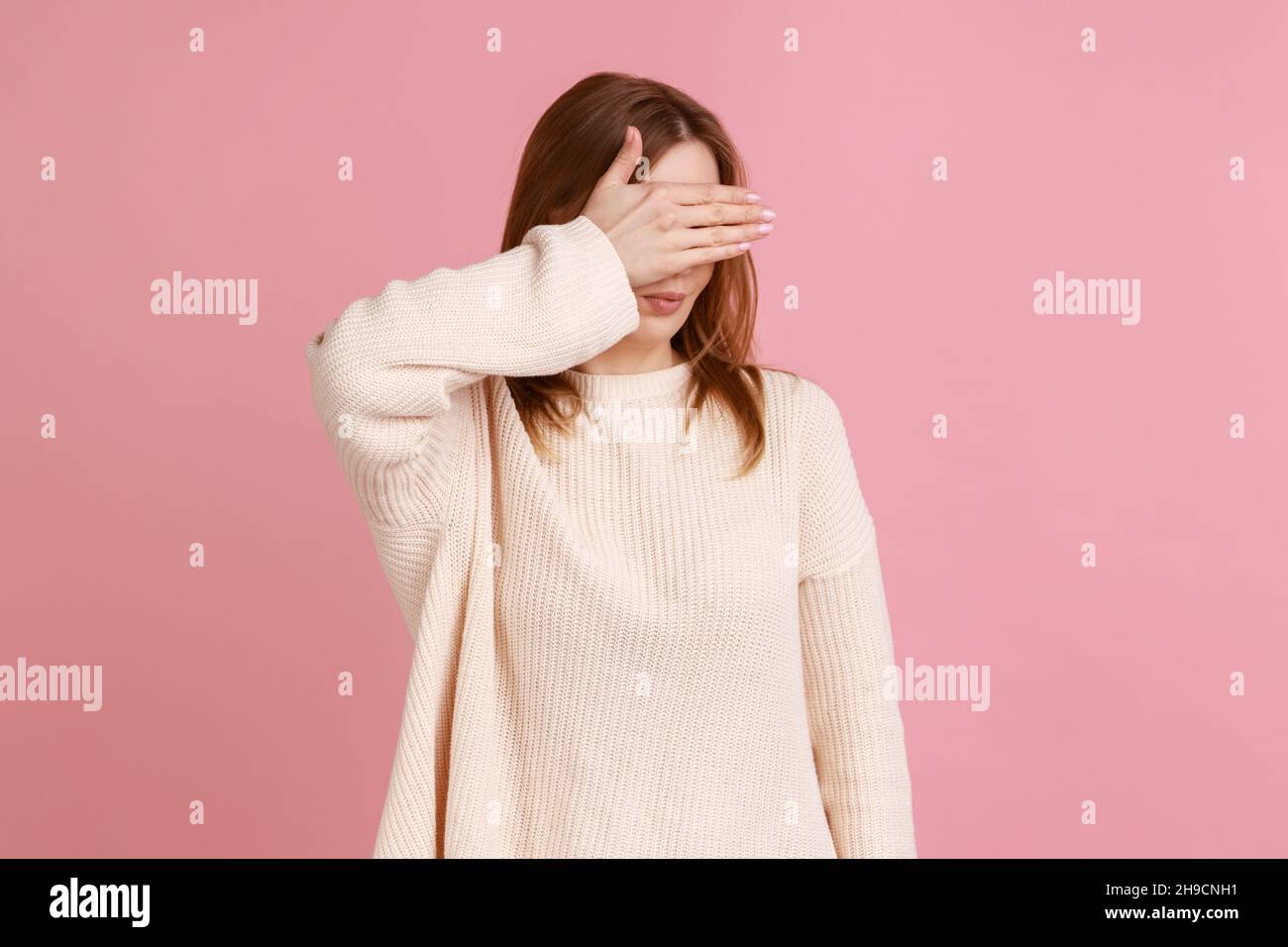 Portrait of ashamed confused attractive blond woman closing eyes with hand, hiding, ignoring dont want to see, wearing white sweater. Indoor studio shot isolated on pink background. Stock Photo