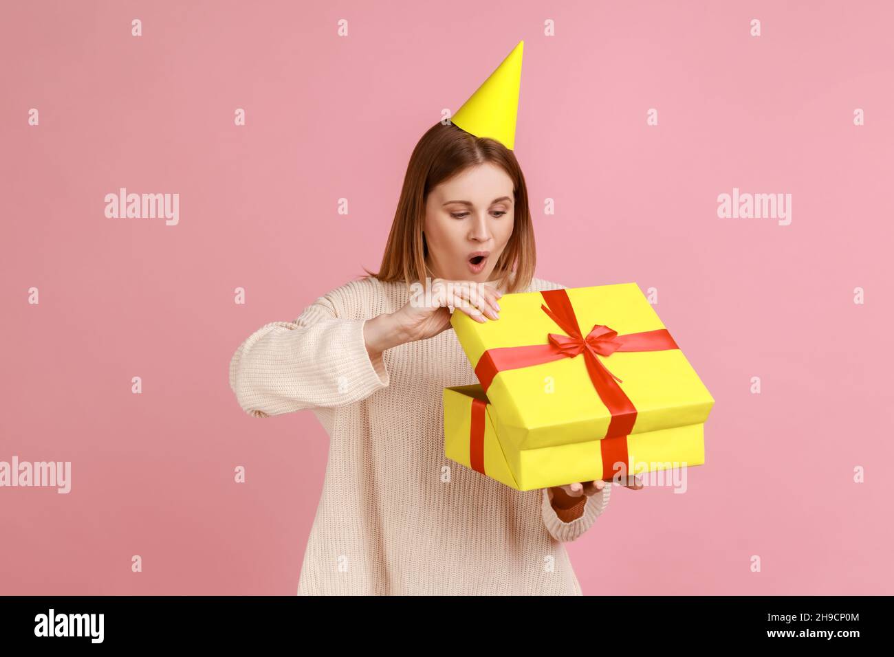 Portrait of excited amazed blond woman in party cone unpacking present box, having pleasant surprise, being shocked, wearing white sweater. Indoor studio shot isolated on pink background. Stock Photo