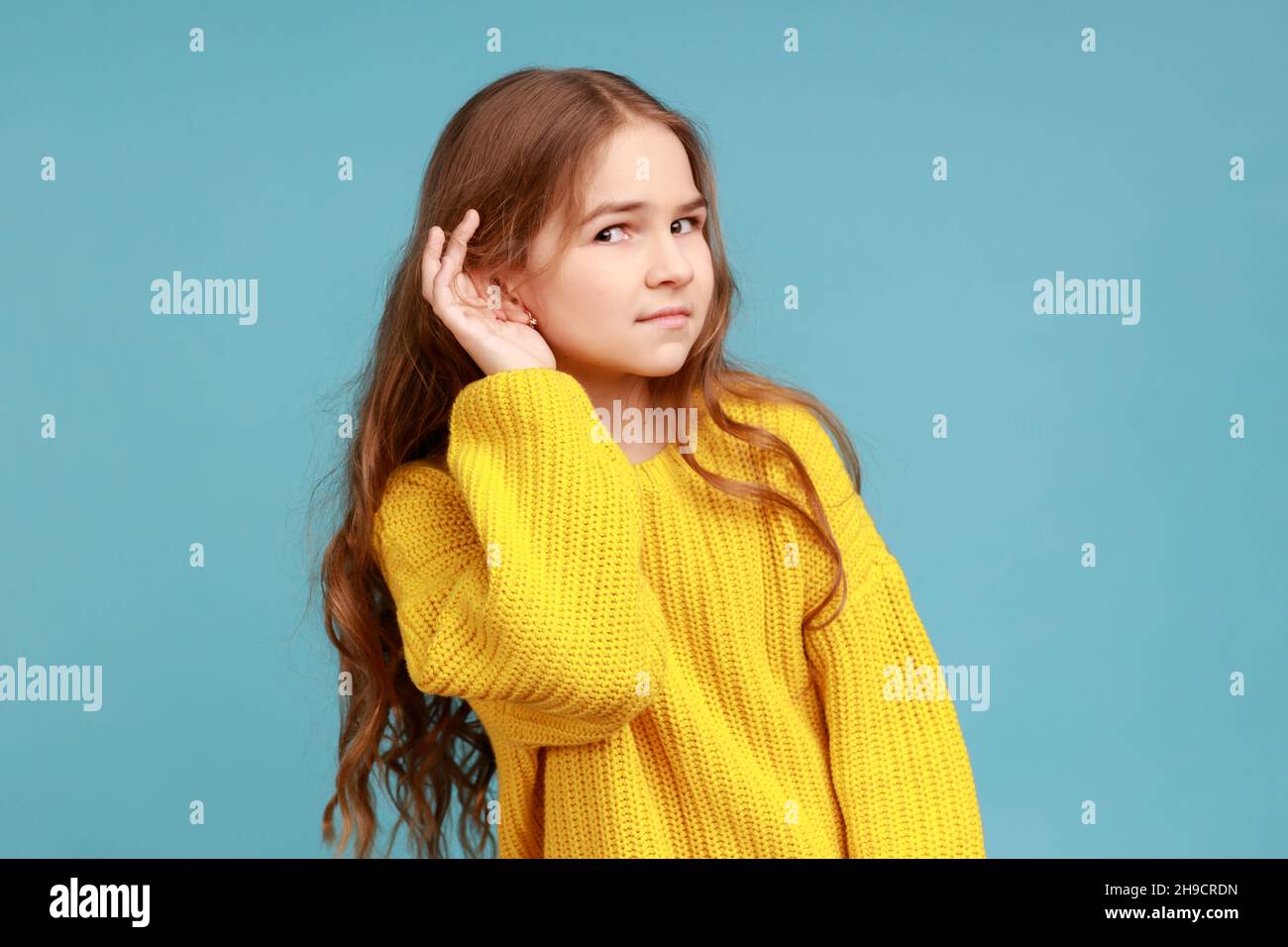 Portrait of little girl holding hand near ear and listening carefully intently to what you say, wearing yellow casual style sweater. Indoor studio shot isolated on blue background. Stock Photo