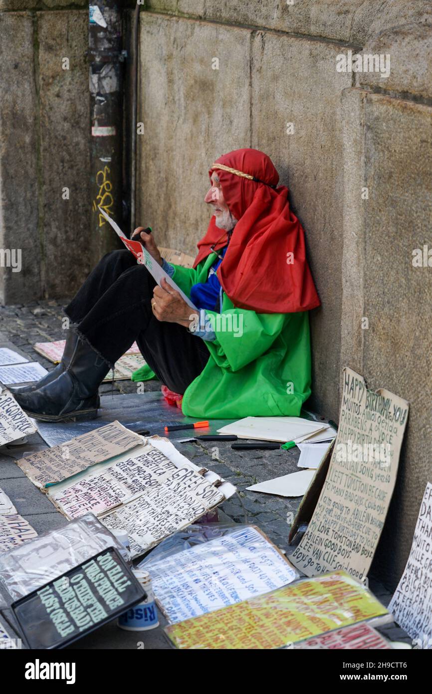 A man with a red headscarf and green jacket sits on the ground of a sidewalk and draws greeting cards that he sells to passersby. Stock Photo