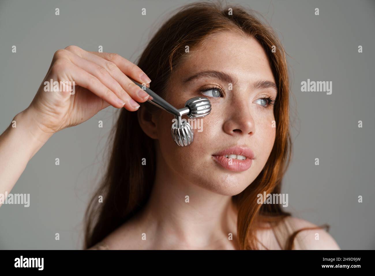 Young white woman using face massager while standing isolated on gray background Stock Photo