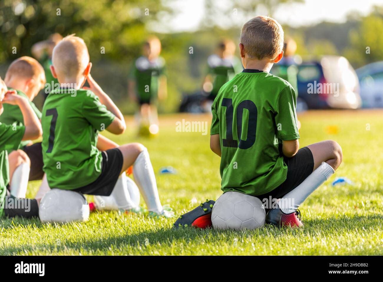 Group of Young Boys Kicking Sports Soccer Game on Grass Pitch. Boys Sitting on Soccer Balls Waiting to Play the Game. Friends in Sports Team. School B Stock Photo