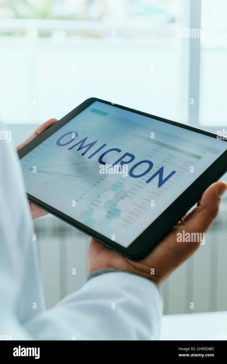 a doctor man, wearing a white coat, has a digital tablet in his hands with the text omicron in its screen, for the SARS-CoV-2 Omicron variant Stock Photo