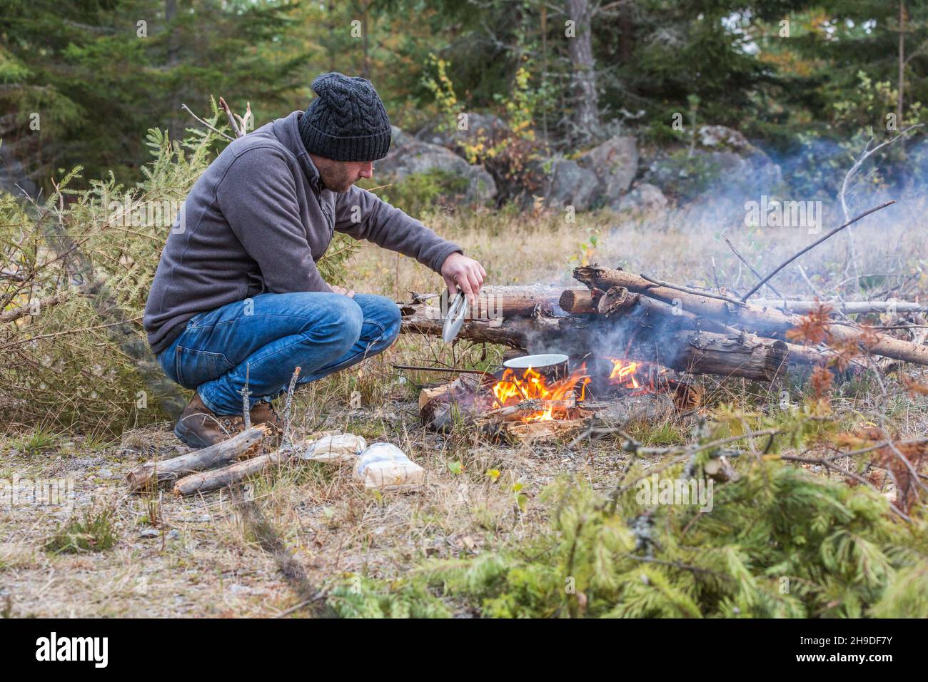 An adventurous man dressed in warm clothing and woolen hat cooks food on a smoking wood-fired campfire with lots of ready firewood to make a fire Stock Photo