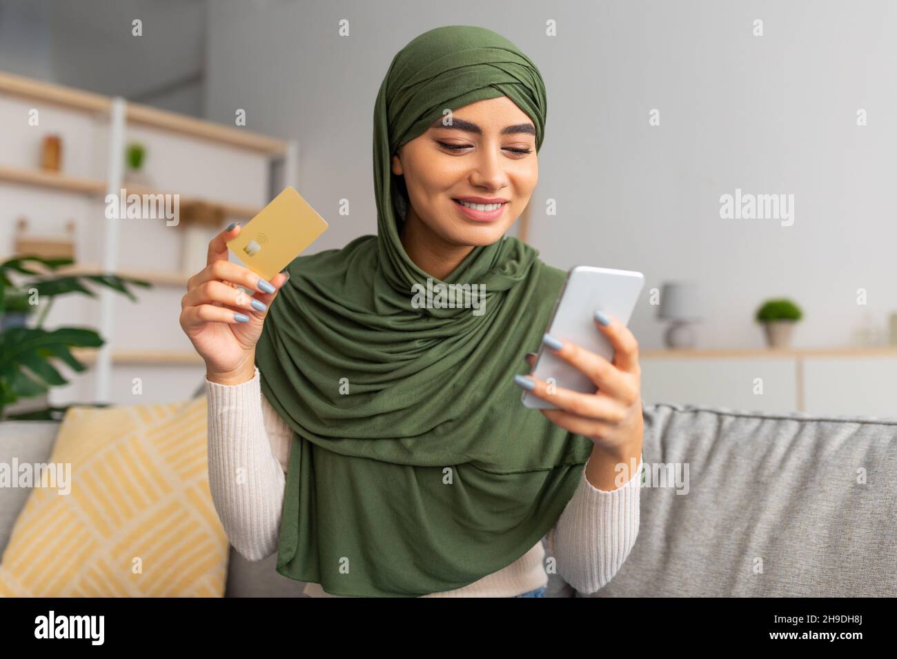 Happy Arab lady in hijab sitting on sofa with smartphone and credit ...