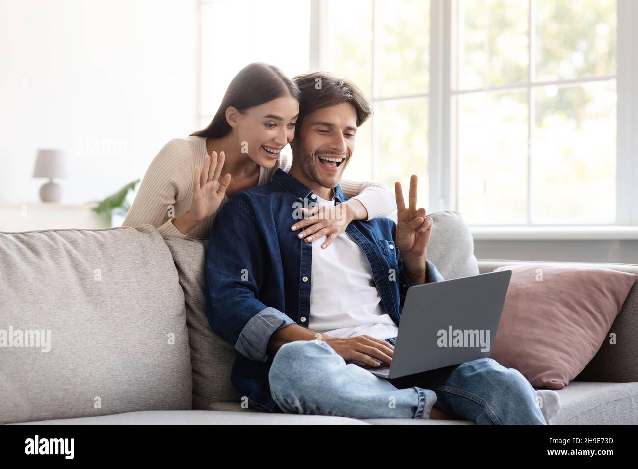Smiling millennial european couple communicate with family remotely, wave hand, showing peace sign Stock Photo