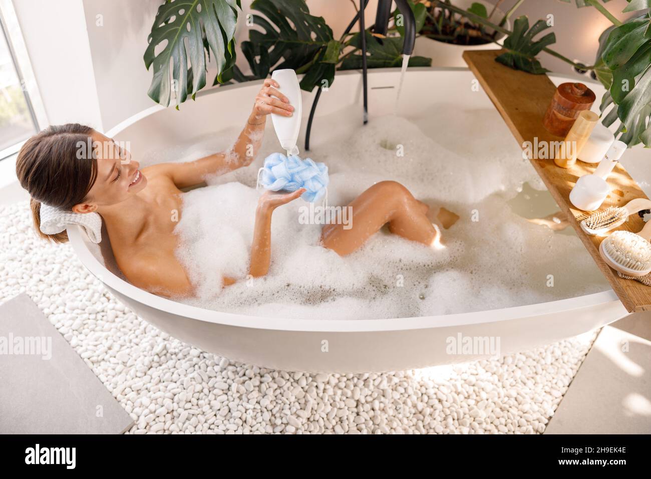 Relaxed young woman pouring shower gel from bottle onto loofah sponge while bathing Stock Photo